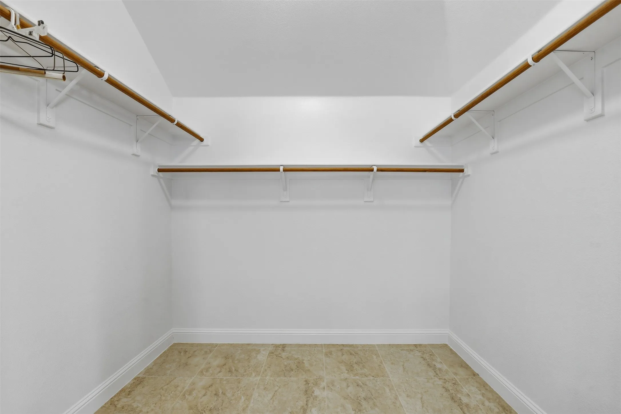 Spacious walk-in closet featuring light-toned tile flooring, white baseboards, and multiple wood rods with white shelving brackets for ample storage