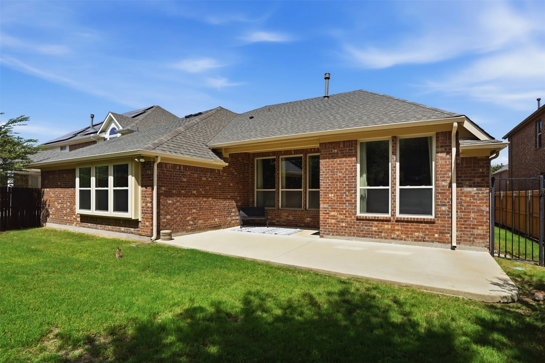 Expansive concrete patio with a brick exterior, multiple windows with white trim, a shingled roof, and a green lawn