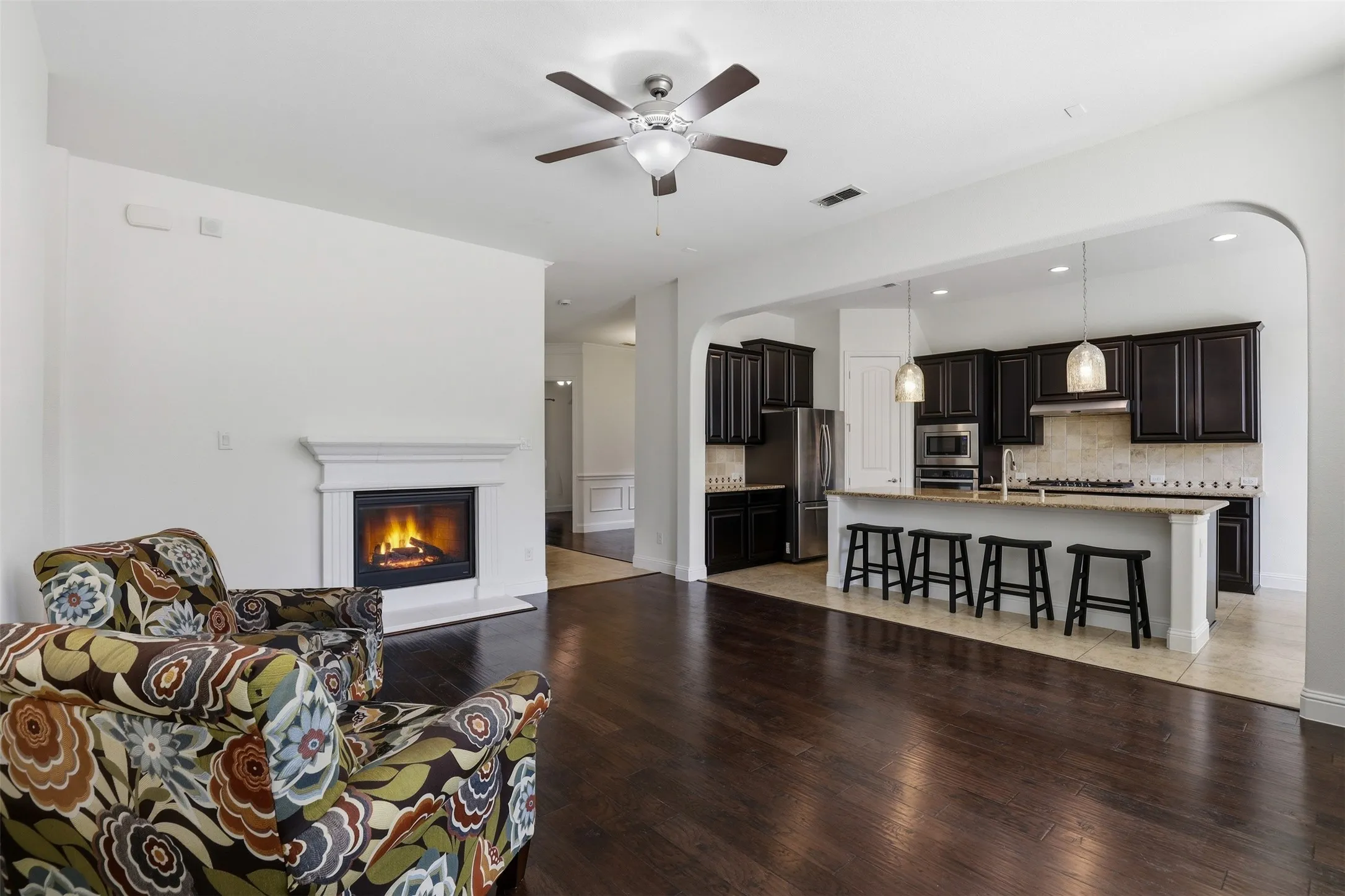 Open-concept living area featuring dark wood-finish flooring, a white mantel fireplace, and a ceiling fan