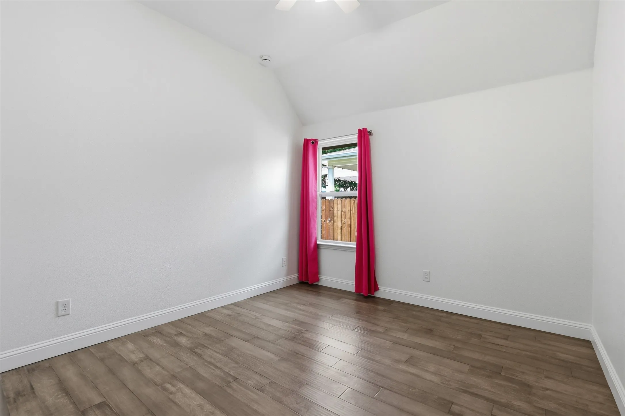 Spacious room featuring vaulted ceilings, gray wood-finish flooring, a window with a white frame, and a white ceiling fan