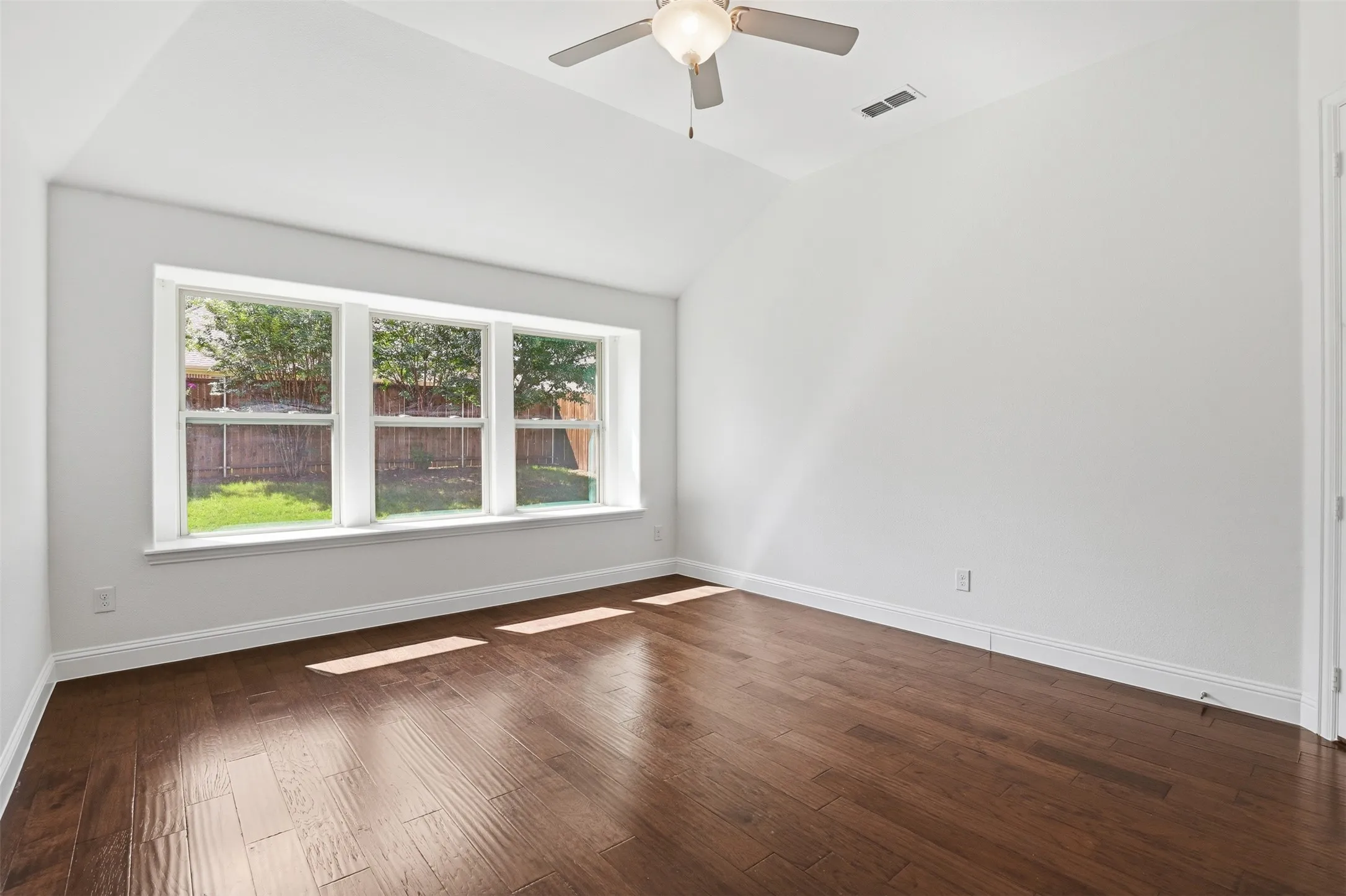 Spacious room featuring rich wood-finish flooring, a contemporary ceiling fan, and abundant natural light from a triple window arrangement