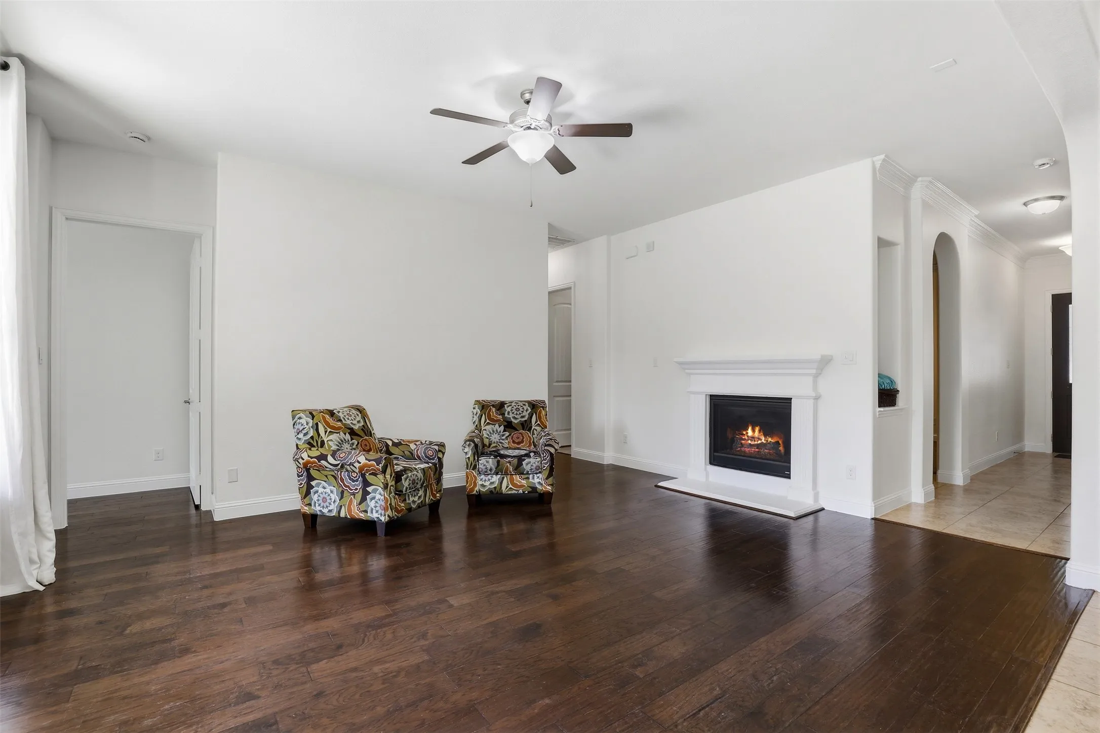 Spacious living area featuring rich wood-finish flooring, a white mantel fireplace, a ceiling fan with integrated lighting