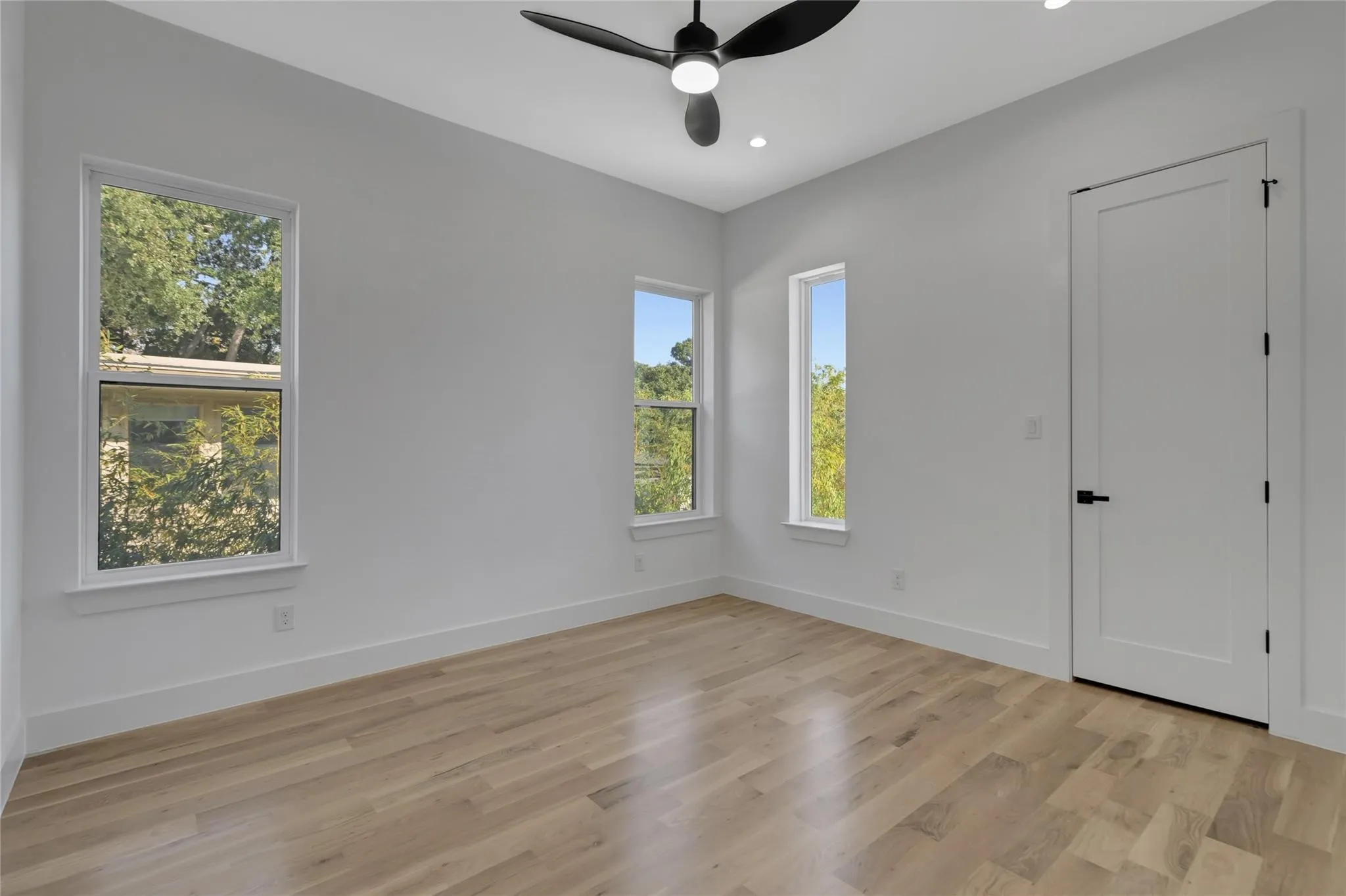 Spacious room featuring wood-finish flooring, multiple windows, a modern ceiling fan with integrated lighting, recessed ceiling lights, and a panel door with matte black hardware
