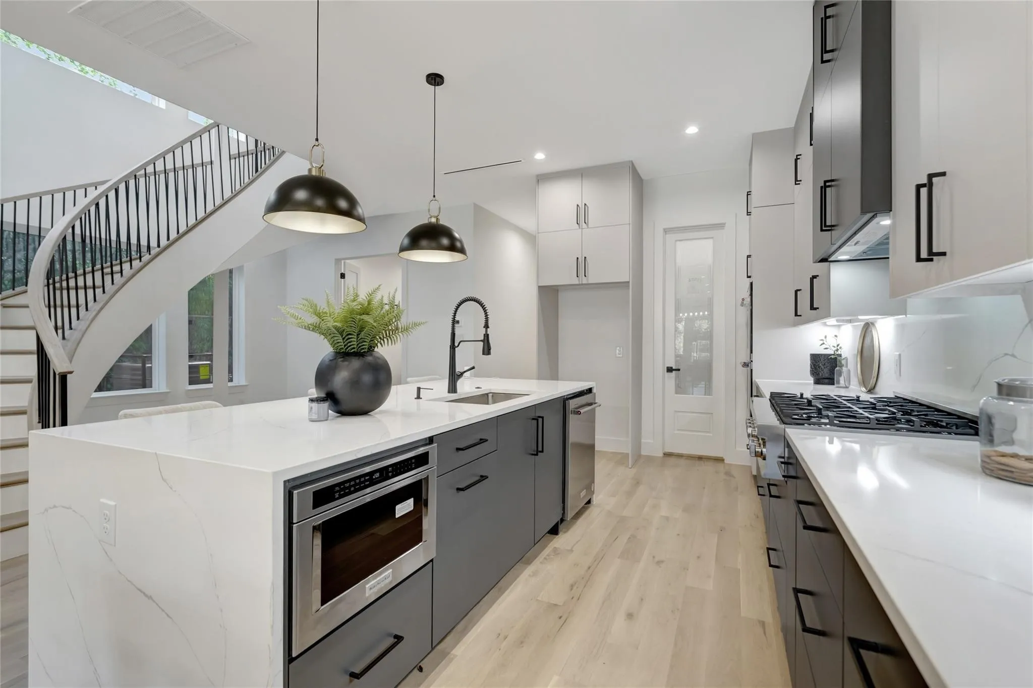 Modern kitchen featuring a waterfall island with integrated microwave, stainless steel appliances, light wood-finish flooring, and a curved staircase with black metal balusters
