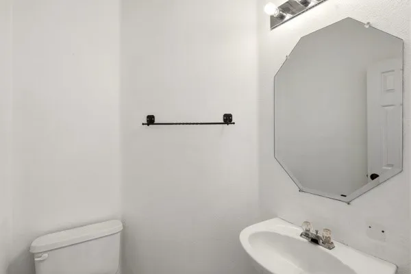 Bathroom featuring an octagonal wall-mounted mirror, vanity lighting, wall-mounted sink with faucet, white toilet, and a dark-toned towel bar