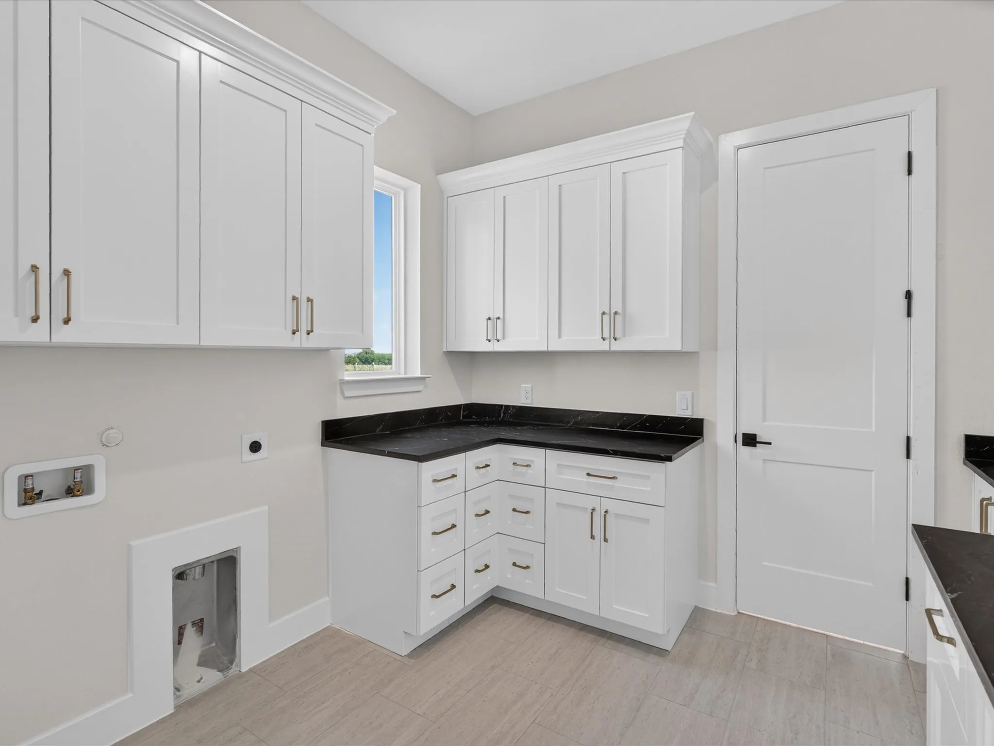 White shaker-style cabinetry with gold-tone hardware, dark countertops, a window, and light-toned tile flooring