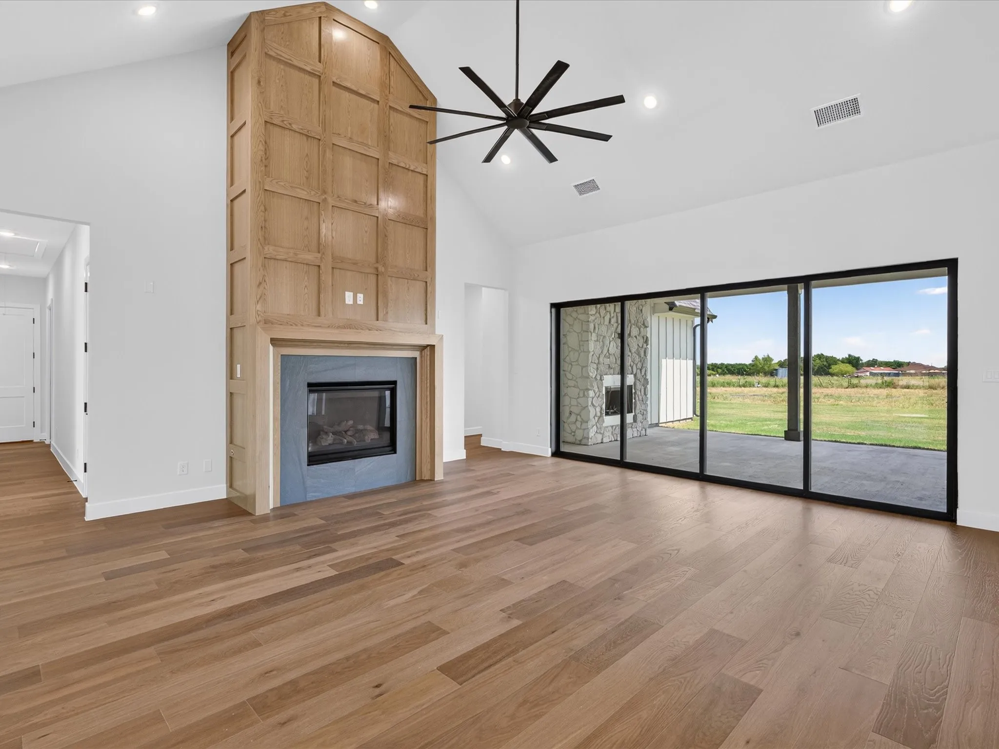 Vaulted ceiling with recessed lighting, wood-finish flooring, fireplace with stone surround, wood-paneled fireplace wall, and black-framed sliding glass doors