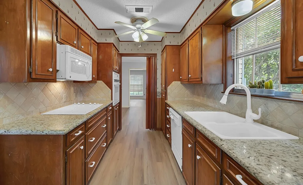 Galley kitchen featuring wood cabinetry, granite-finish countertops, and a full suite of white appliances including a built-in oven, microwave, and dishwasher
