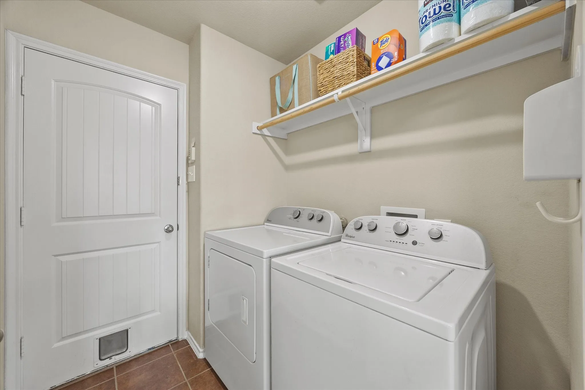 Dedicated laundry area featuring a white door with beadboard paneling, brown floor tiles, and a wall-mounted shelving unit