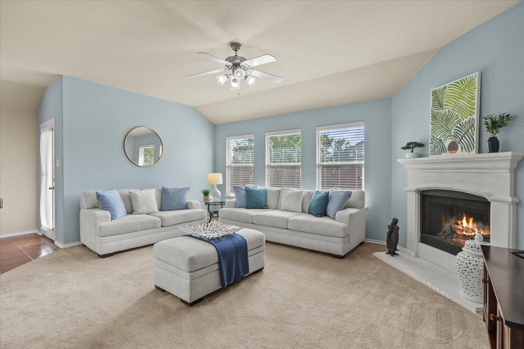 Living area featuring a white mantel fireplace, three windows with horizontal blinds, and a ceiling fan with integrated lighting
