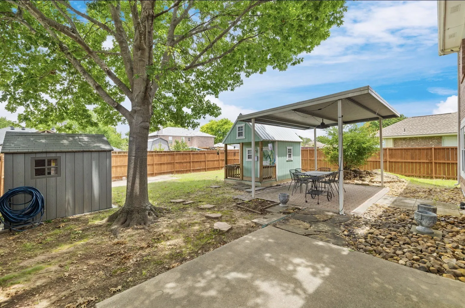Expansive backyard featuring a large tree, a storage shed, a separate small structure with a porch, a covered patio with a paved surface, and a concrete patio