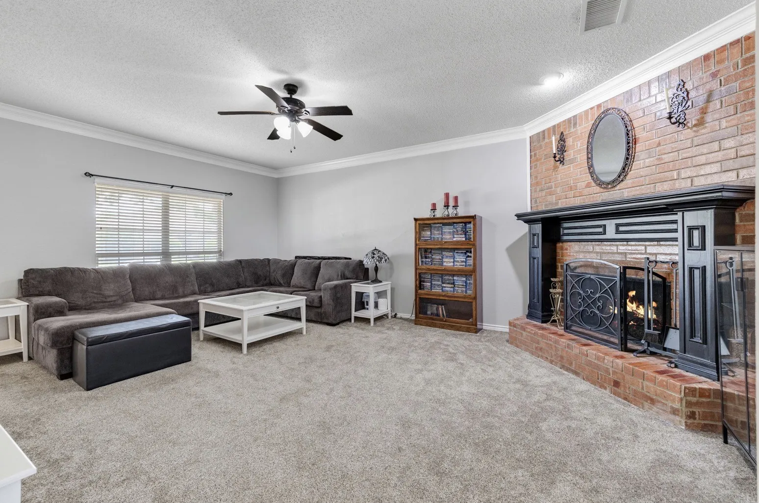 Spacious living area featuring a brick fireplace with a dark wood mantel, grey wall paint, crown molding, and a ceiling fan with integrated lighting