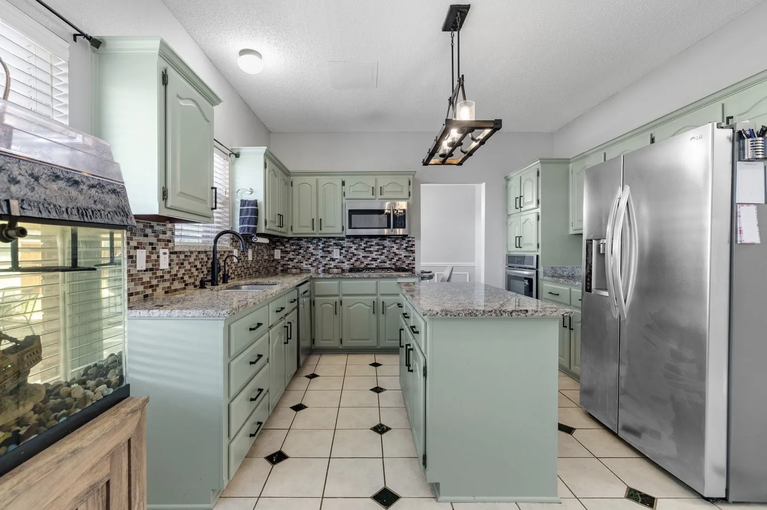 Kitchen featuring light green cabinetry, granite countertops, patterned tile backsplash, stainless steel appliances, and a central island