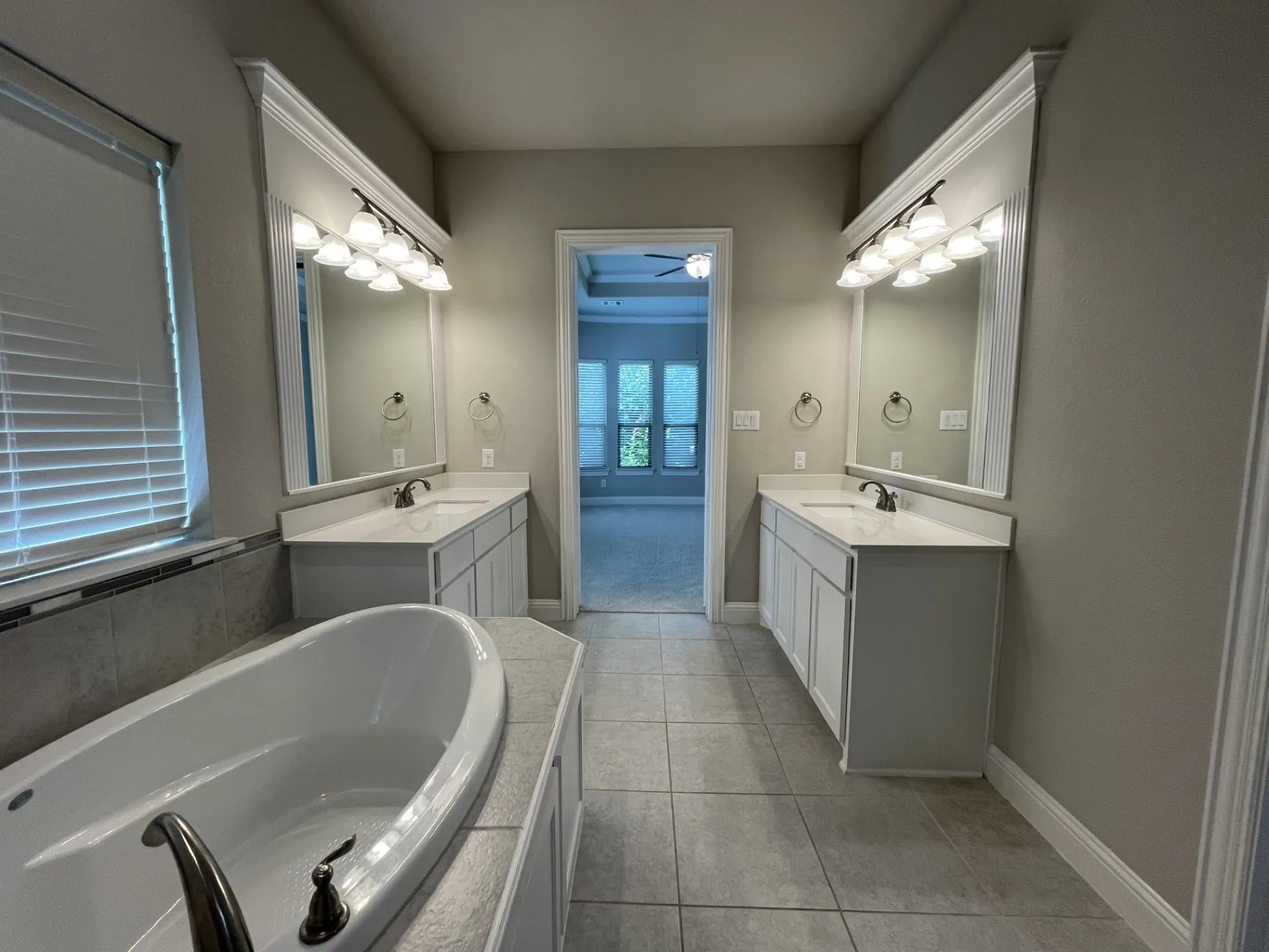 Freestanding soaking tub with tiled surround, dual vanity sinks with white cabinetry and countertops, framed mirrors with overhead lighting, and gray tile flooring