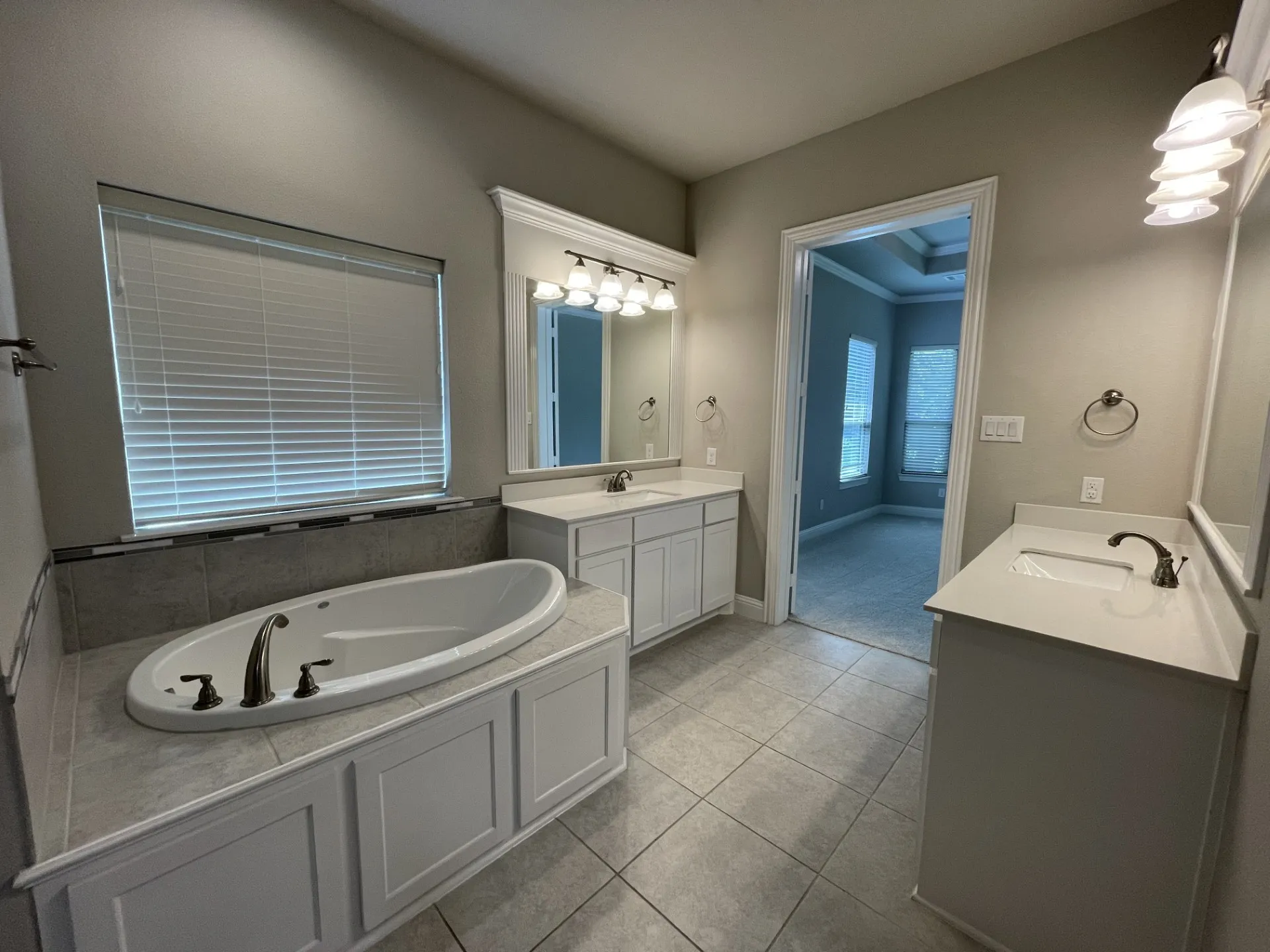 Bathroom featuring a built-in soaking tub with brushed nickel fixtures, dual vanity sinks with light-toned countertops, white shaker-style cabinetry, and tiled flooring