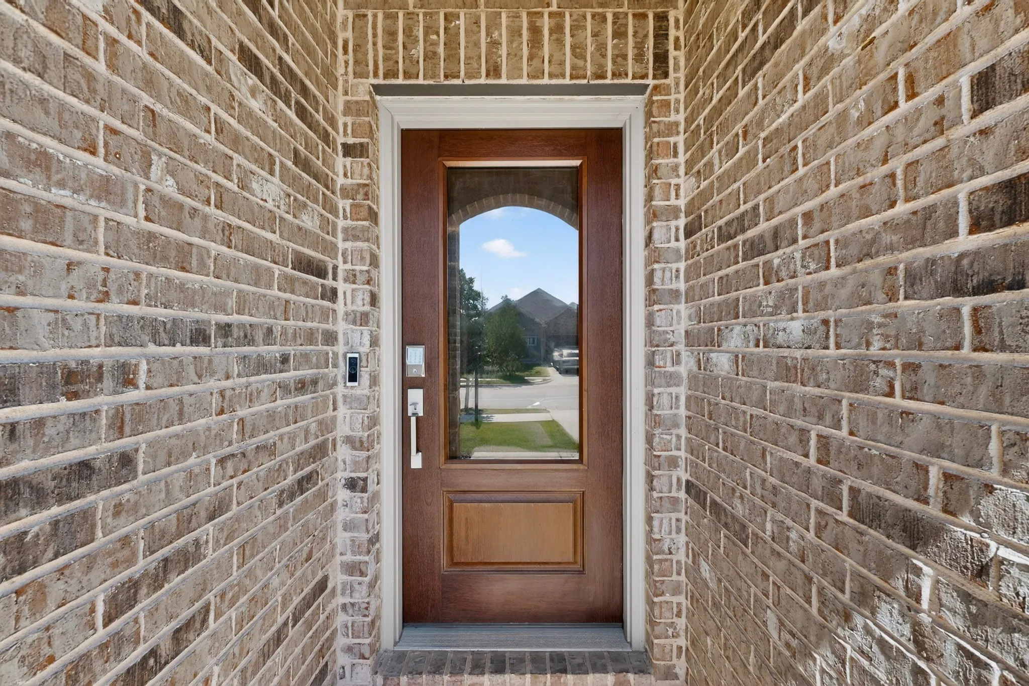 Entryway featuring a wood-finish door with an arched glass panel, a smart doorbell system, and a white trim surround, all framed by a brick facade
