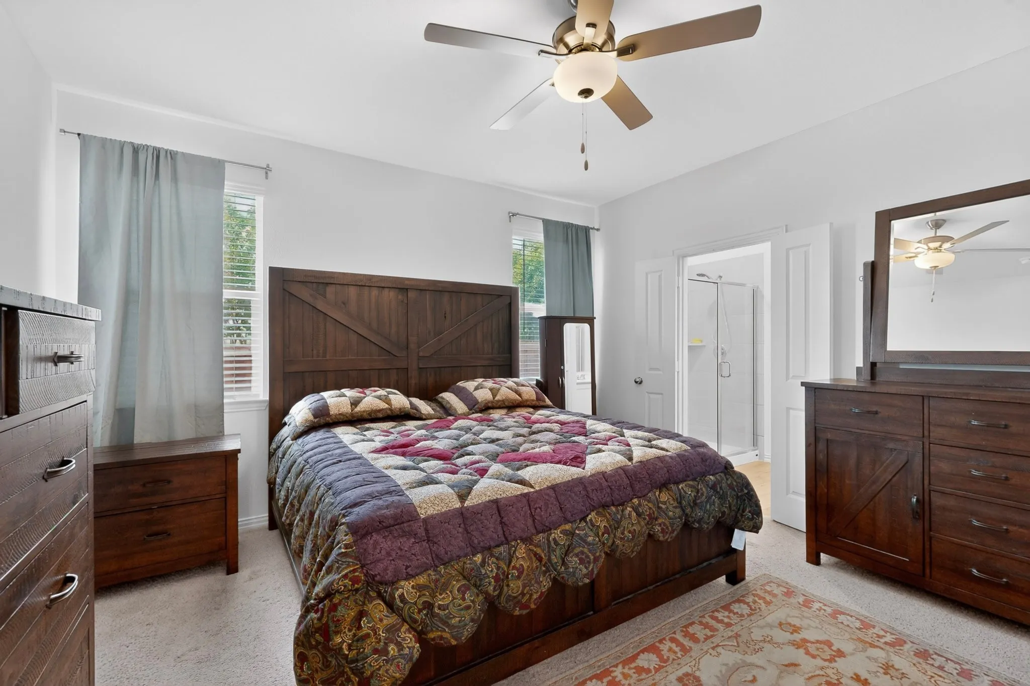 Carpeted bedroom with a ceiling fan light fixture, two windows with blinds, and an open doorway revealing a shower enclosure