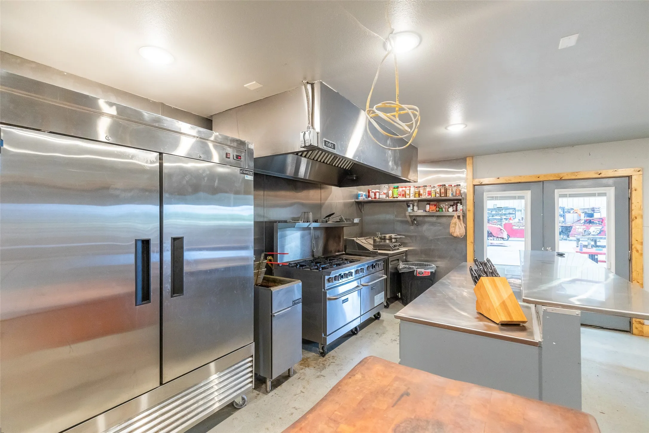 Commercial-grade kitchen featuring a large stainless steel double-door refrigerator, a professional range with an oversized vent hood, and extensive stainless steel countertops