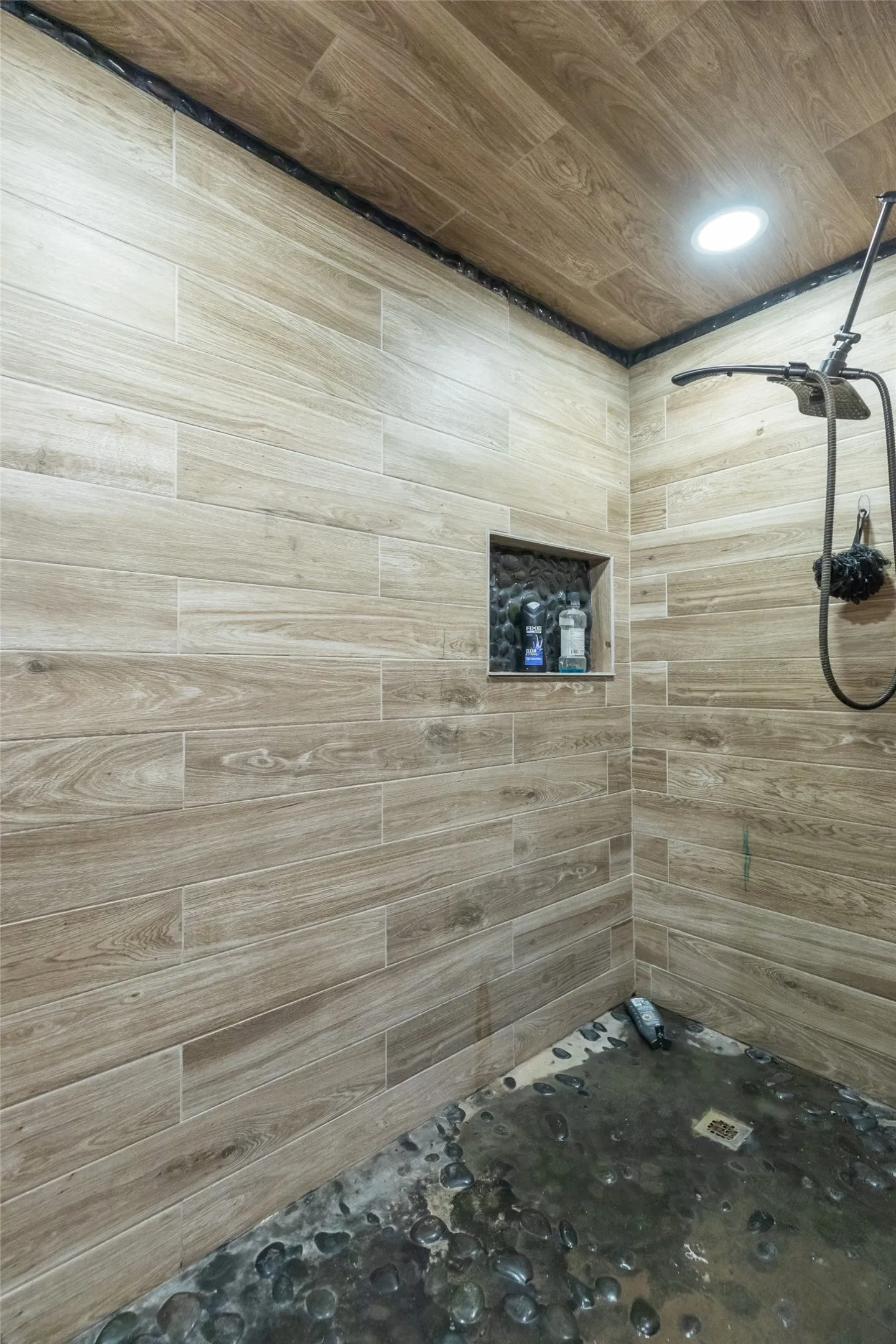 Shower stall featuring wood-finish tile walls and ceiling, a built-in shower niche, a rain showerhead with a separate handheld spray, and a pebble-stone floor