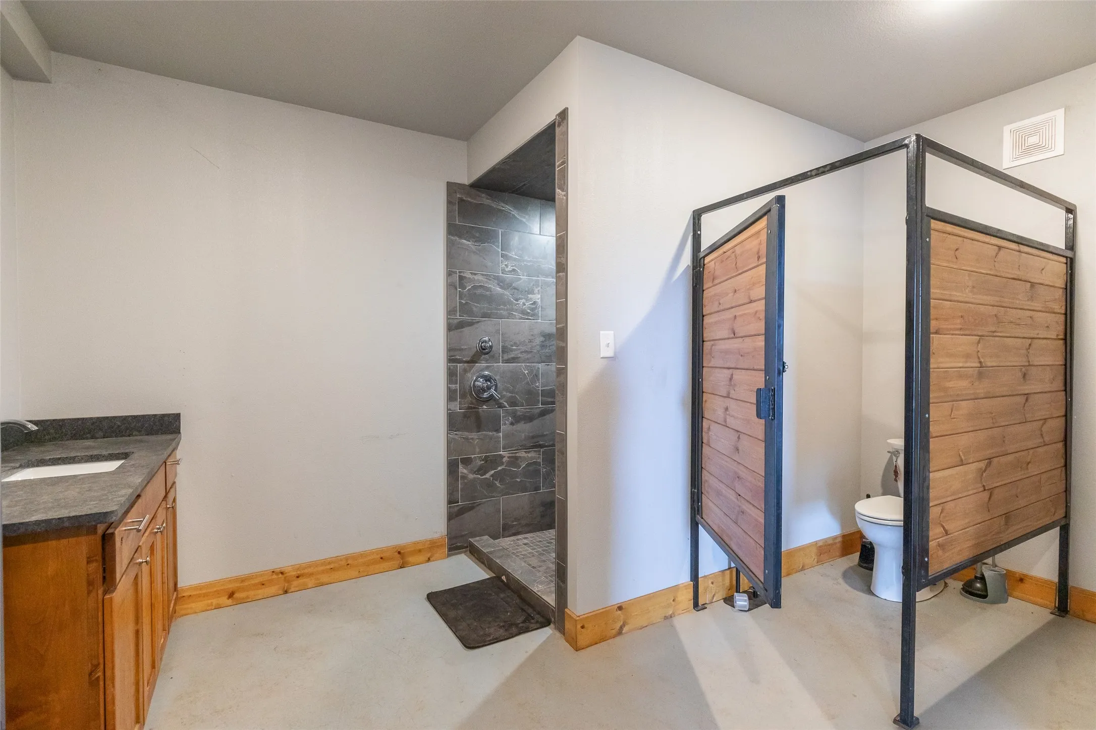 Bathroom featuring a built-in wood vanity with a dark countertop and undermount sink, a tiled shower enclosure, and a toilet with wood-panel privacy walls