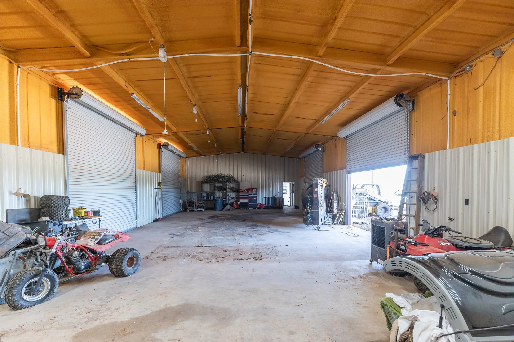 Spacious metal building featuring two large roll-up doors, high ceilings with exposed beams, fluorescent lighting, and a concrete floor