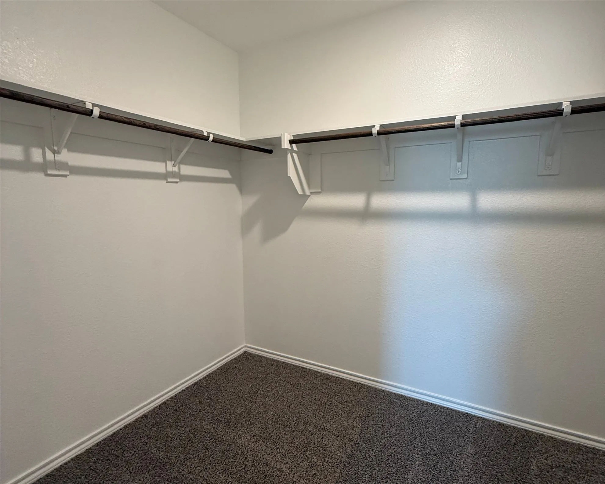 Primary carpeted closet space featuring L-shaped hanging rods and white baseboards