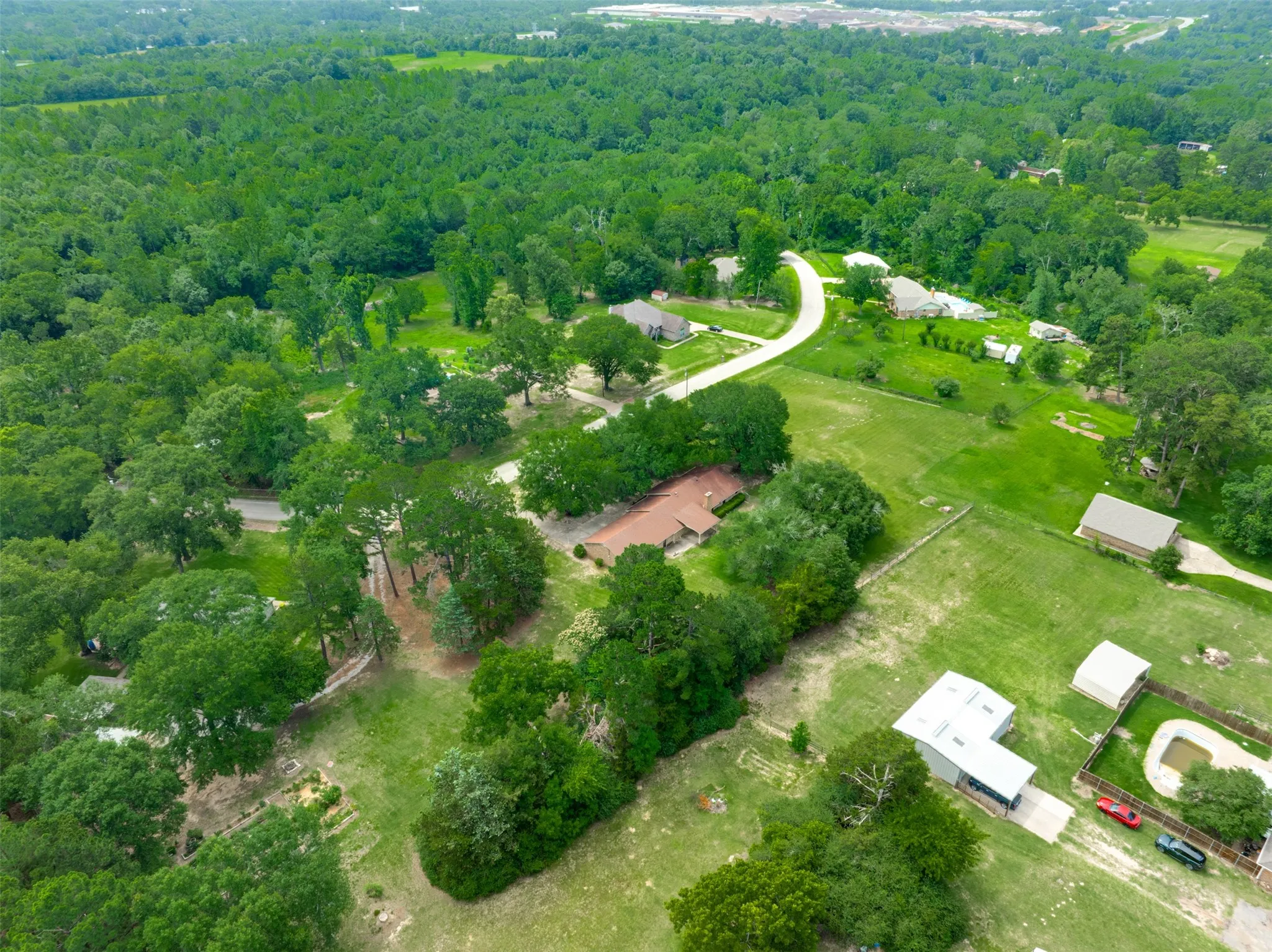 Expansive rural setting featuring a winding paved road, multiple residential properties, dense tree lines, and open green spaces