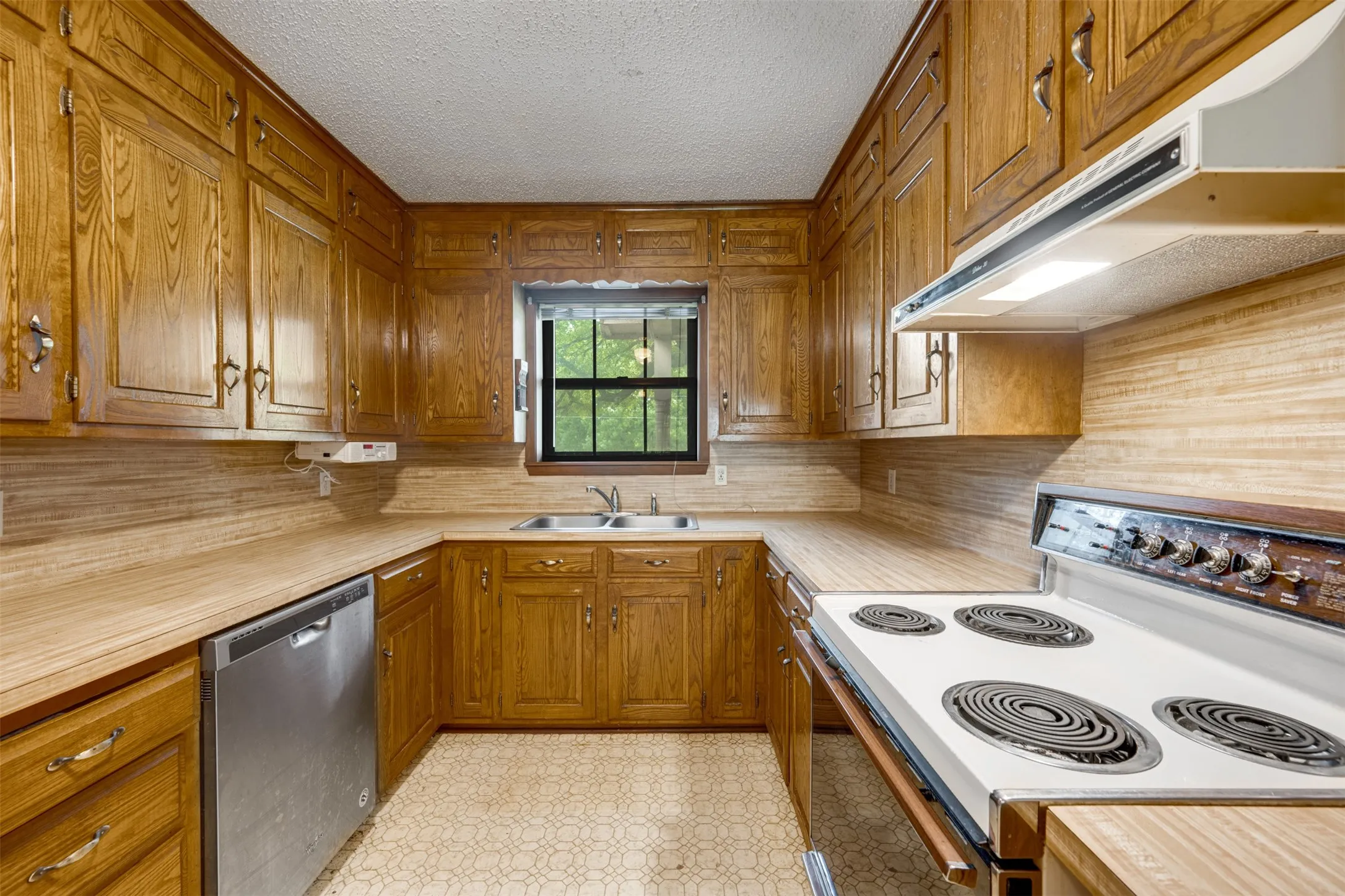 U-shaped kitchen featuring wood cabinetry, laminate countertops, a stainless steel dishwasher, a white electric range with overhead ventilation, and a double basin sink beneath a window