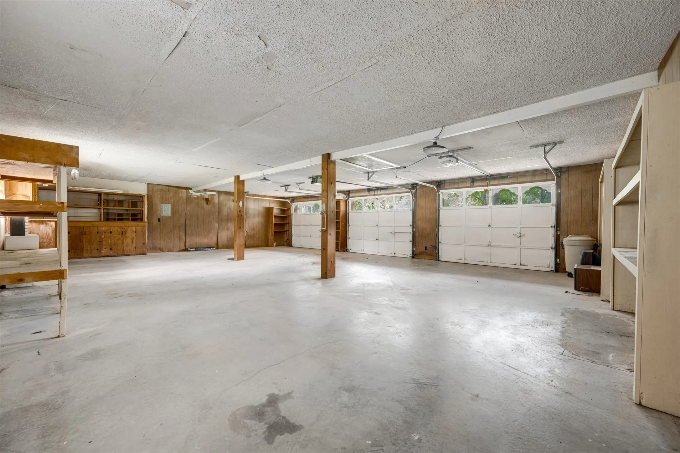 Spacious garage featuring a concrete floor, two white overhead garage doors with window inserts, exposed structural beams, and built-in shelving and cabinetry for storage