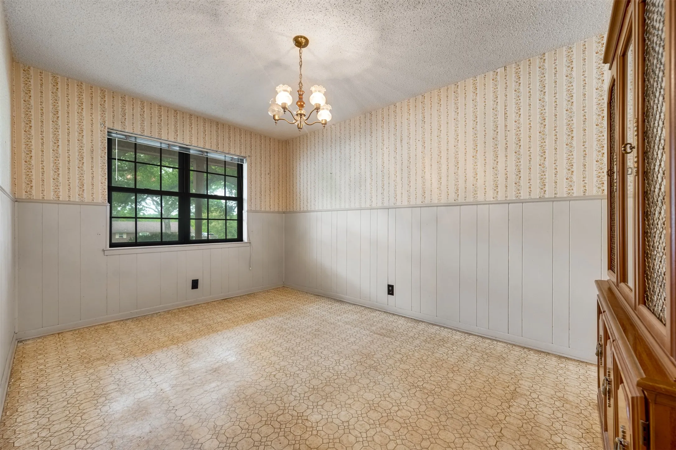 Room featuring a large window with black framing, textured wallpaper, white wainscoting, and a central brass chandelier