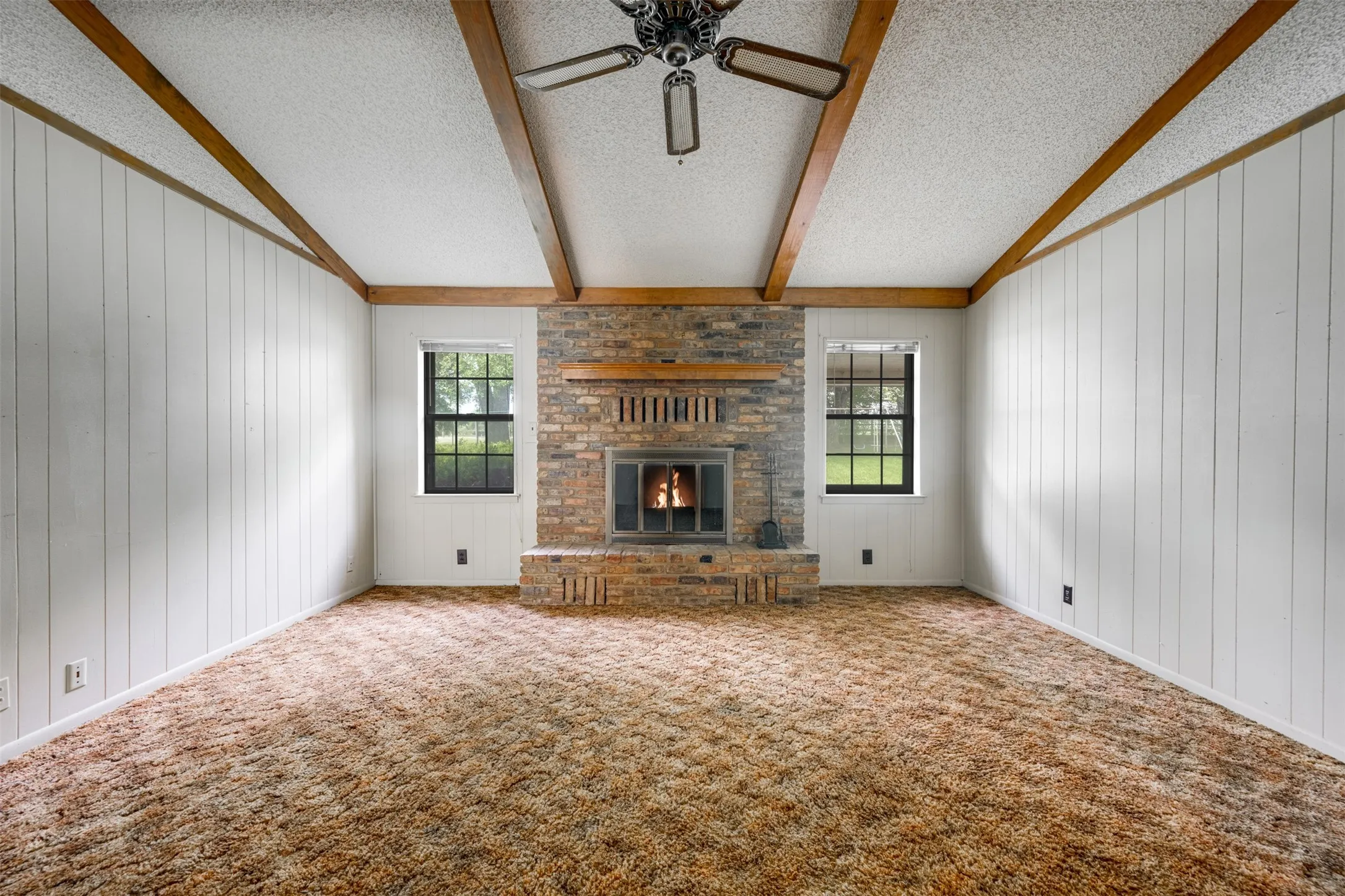 Spacious room featuring a brick fireplace with a wood mantle, exposed wood ceiling beams, white vertical paneling, and two windows with black frames