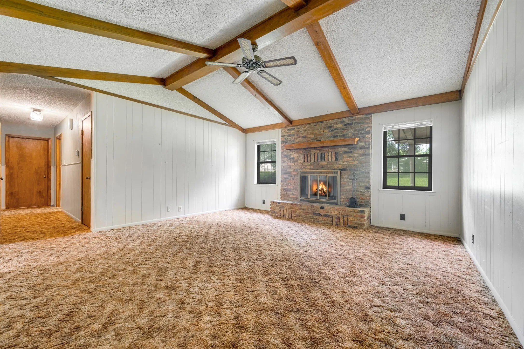 Spacious living area featuring a vaulted ceiling with exposed wood beams, a brick-surround fireplace with a wood mantle, and paneled walls