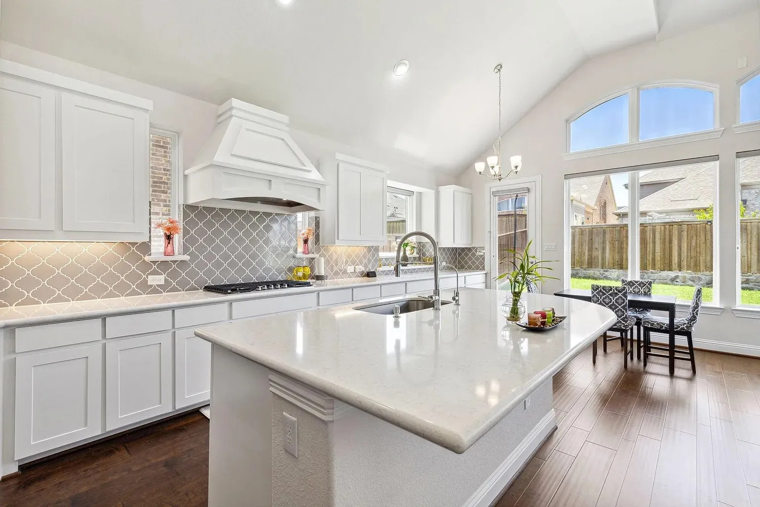 Open-concept kitchen featuring an expansive island with a built-in sink, white shaker cabinetry, a decorative range hood, gray quatrefoil backsplash, and wood-finish flooring