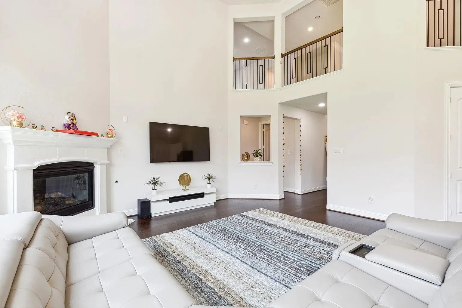 Spacious living area featuring a fireplace with a white mantel, dark wood-finish flooring, high ceilings, and an upper-level walkway with metal railings