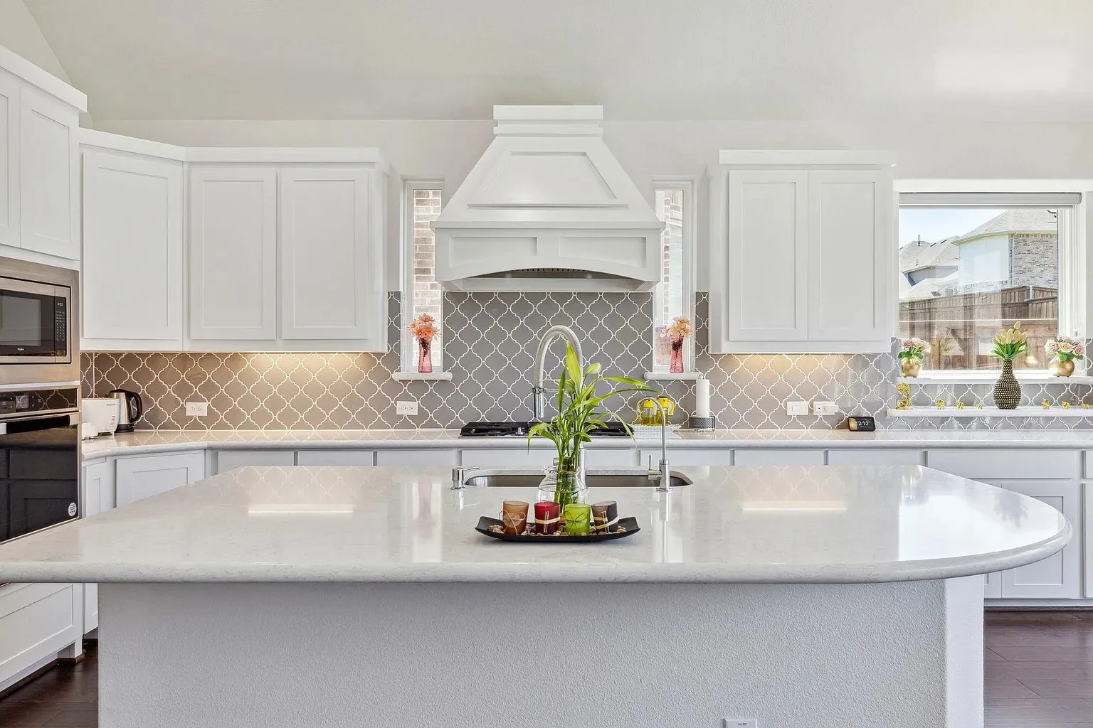 Kitchen featuring an expansive island with a built-in sink, white shaker-style cabinetry, stainless steel appliances, a geometric pattern tile backsplash, and abundant natural light from multiple windows