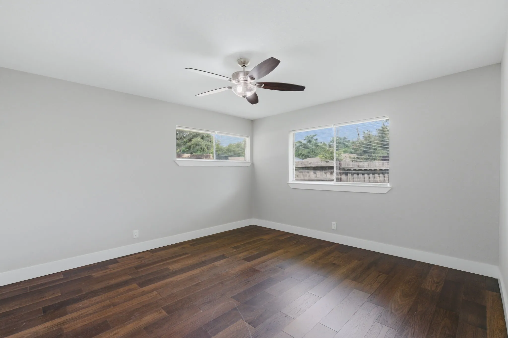Spacious room featuring wood-finish flooring, two large windows, a ceiling fan with integrated lighting, clean white baseboards, and light gray wall paint