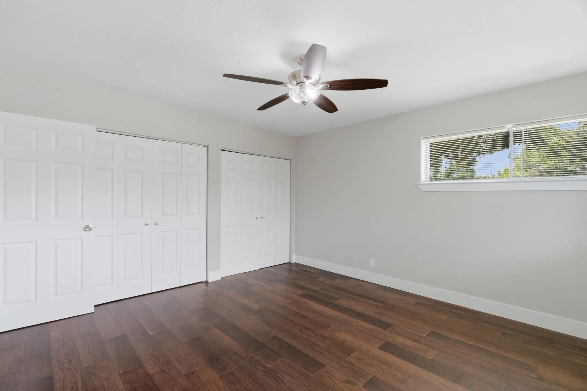 Spacious room featuring wood-finish flooring, two sets of bi-fold closet doors, a ceiling fan with light fixture, and a horizontal window with blinds