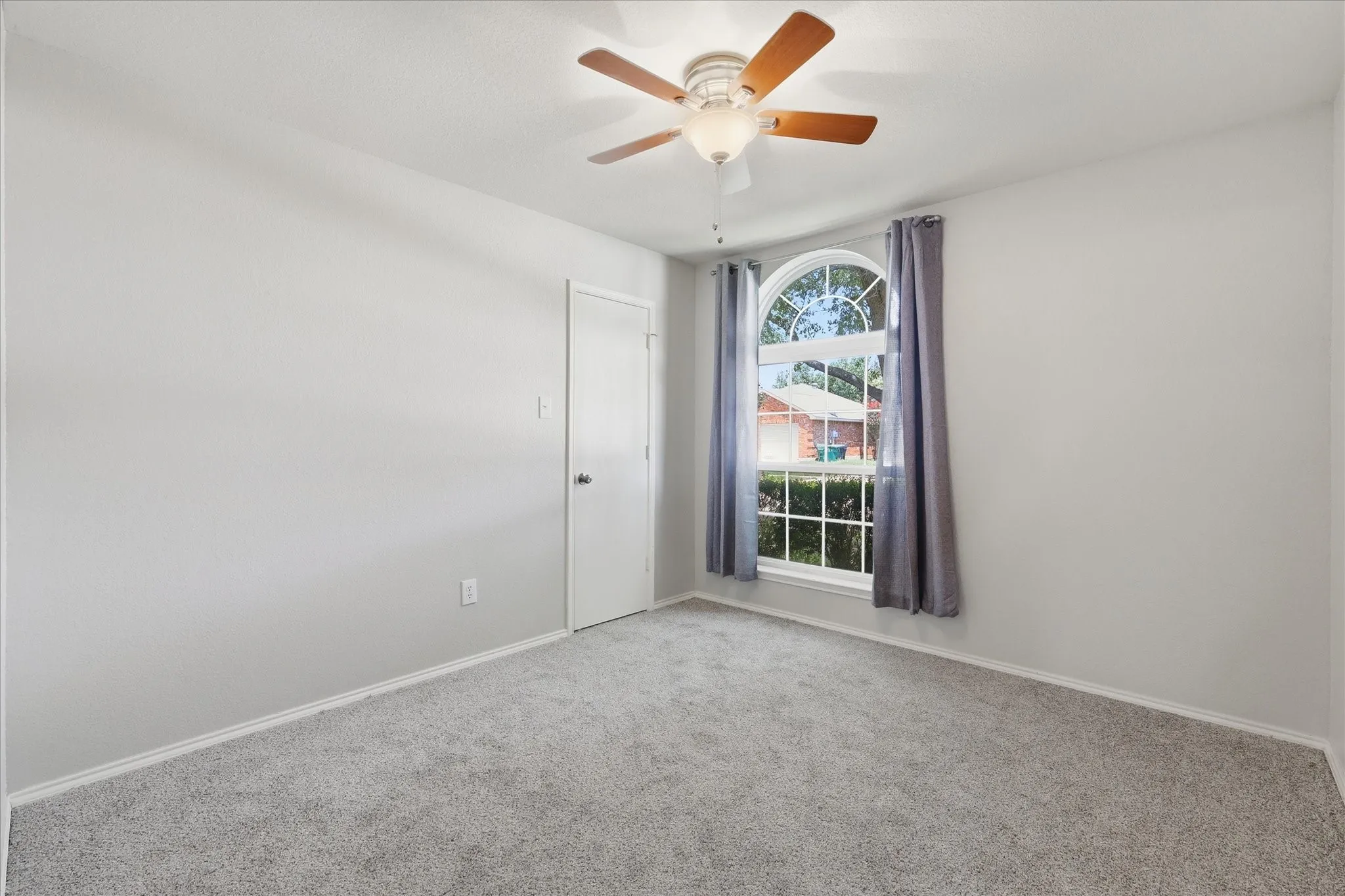 Light gray carpeting with baseboards, a white painted door, a wood-finish ceiling fan with integrated lighting, and an arched window with white trim