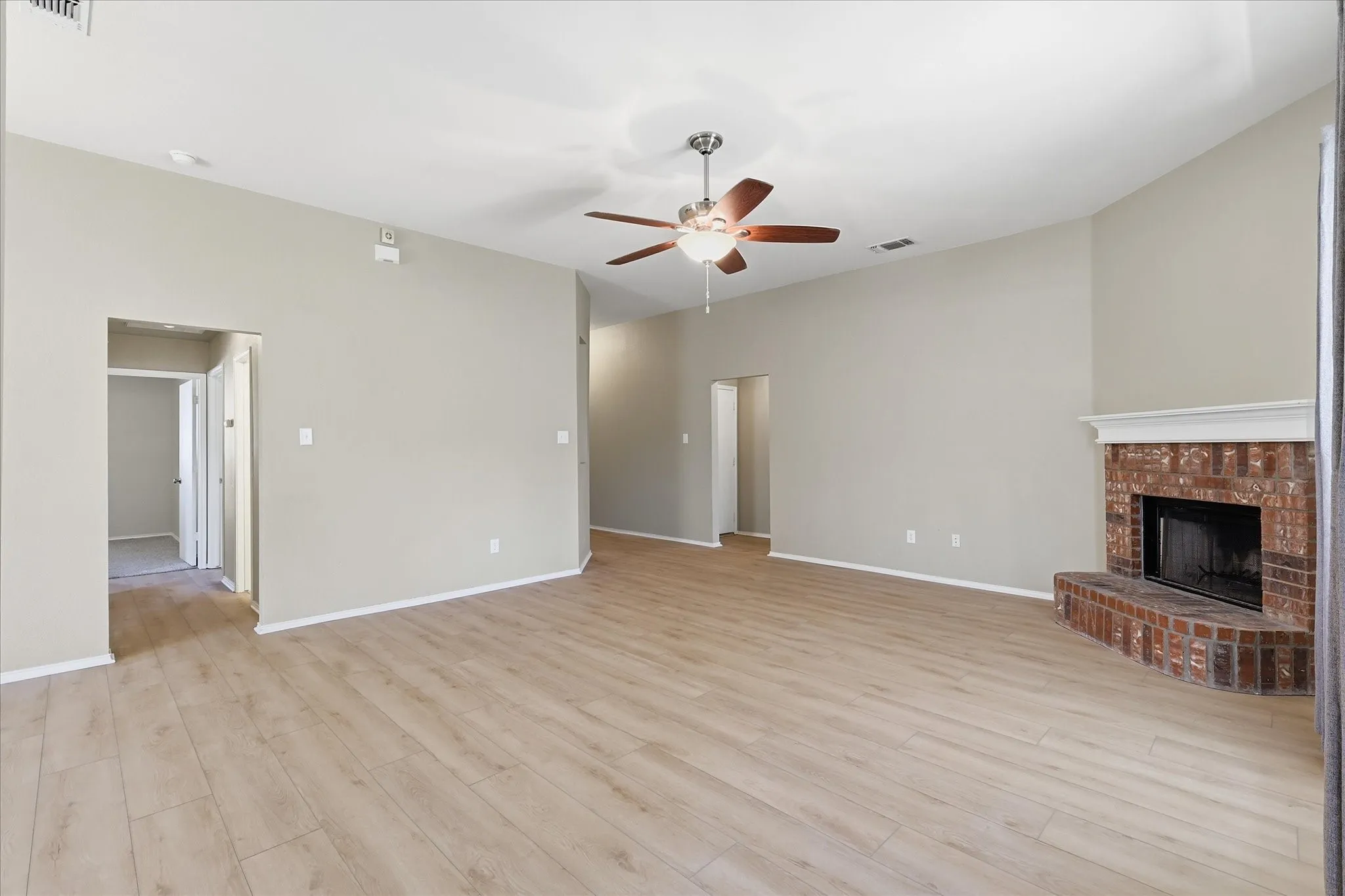 Spacious living area featuring light wood-finish flooring, a brick fireplace with a white mantel, and a ceiling fan with integrated lighting