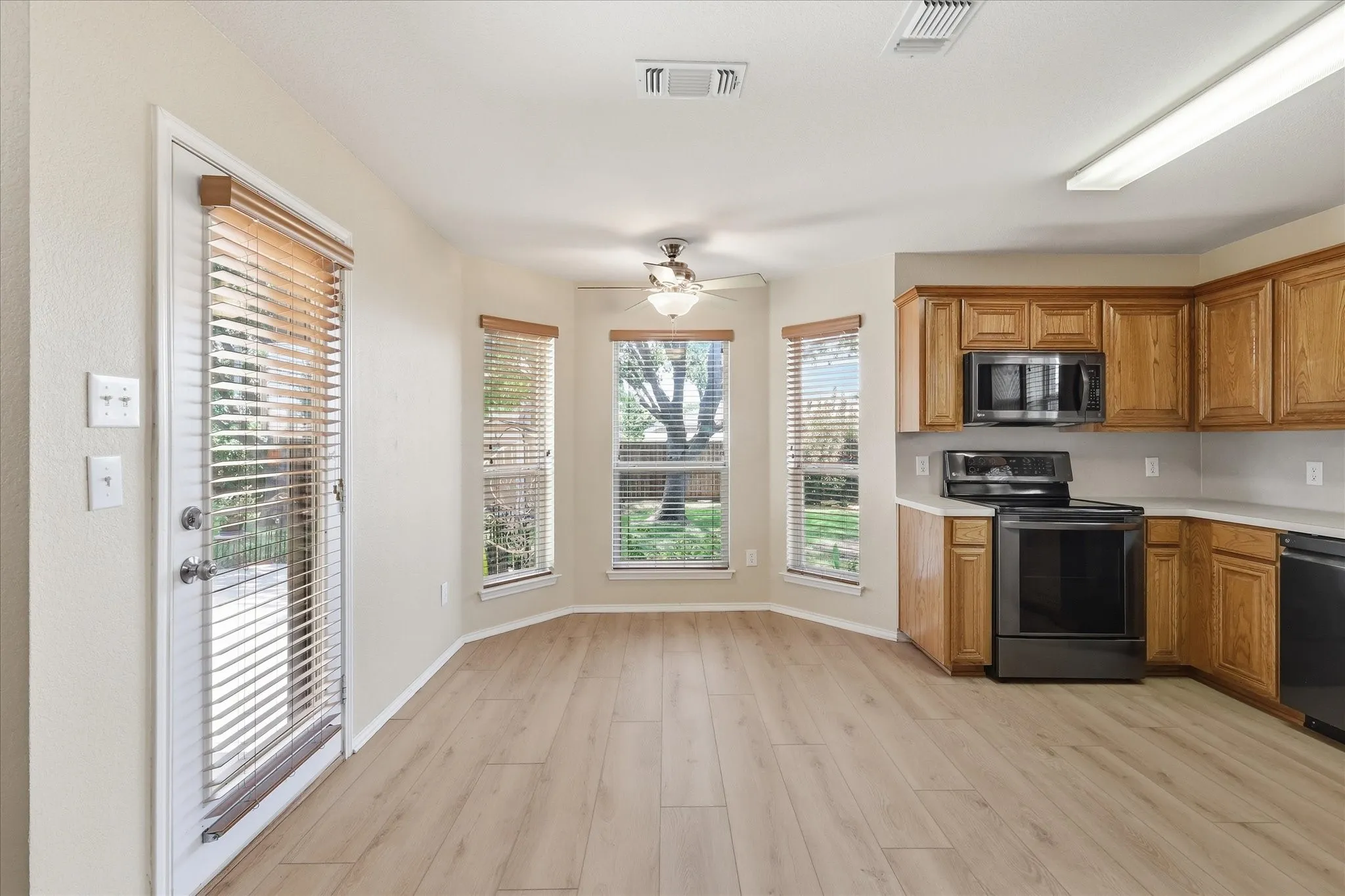 Kitchen and dining area featuring wood-finish flooring, a bay window with blinds, a ceiling fan, light wood cabinetry, and black appliances including a range and microwave