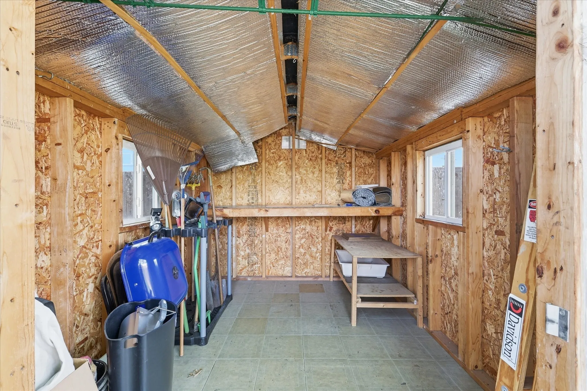 Utility shed interior featuring exposed wood framing, insulated ceiling panels, two windows, built-in shelving, and light-toned floor tiling