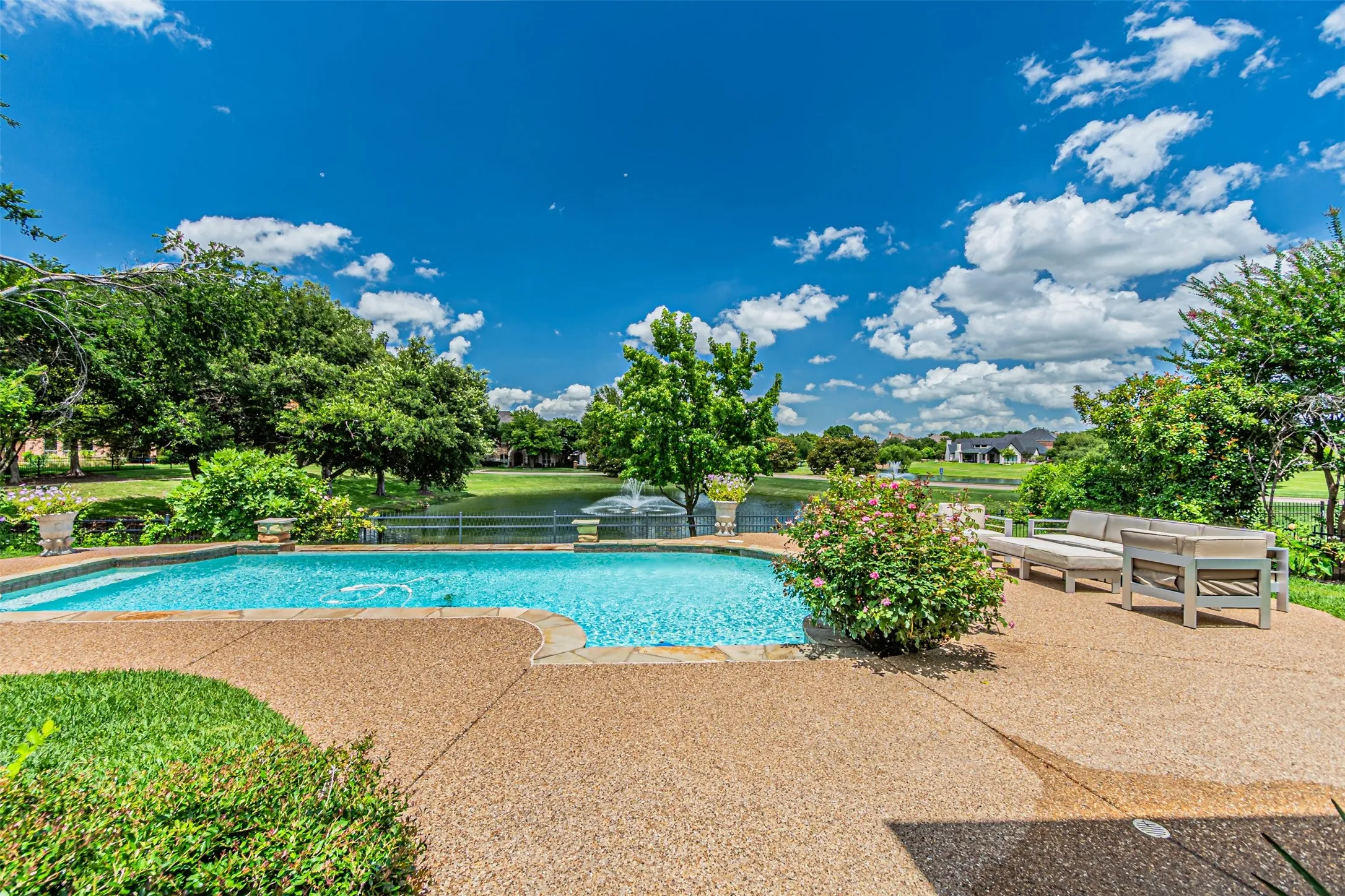 Expansive backyard featuring a sparkling in-ground pool, a spacious aggregate patio, mature landscaping, and a water feature with a pond backdrop
