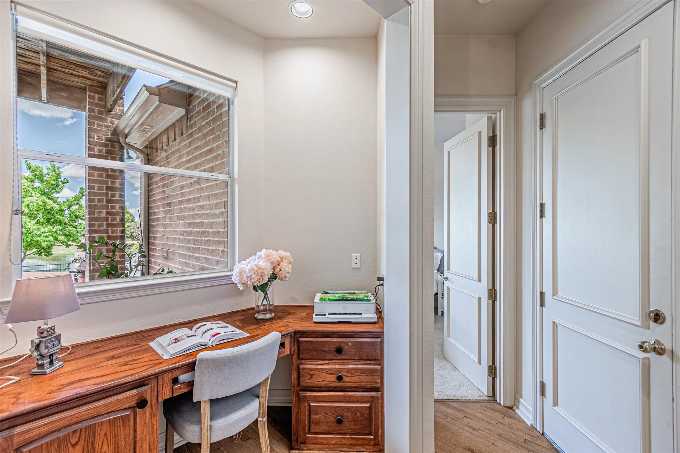 Built-in wood-finish desk with storage drawers, complemented by a large window overlooking a brick exterior and greenery