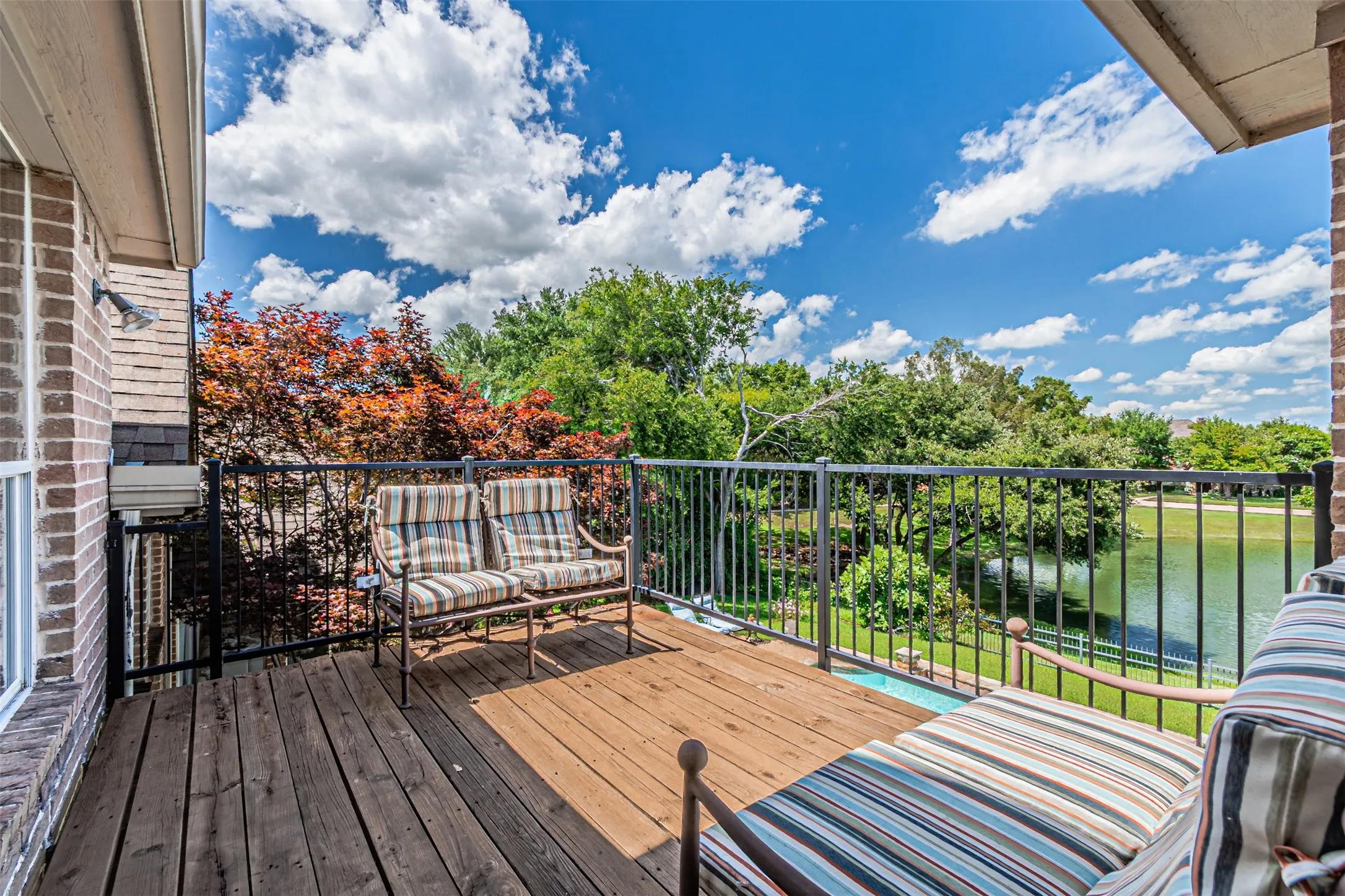 Exterior balcony featuring wood-finish decking, black metal railings, and a brick facade