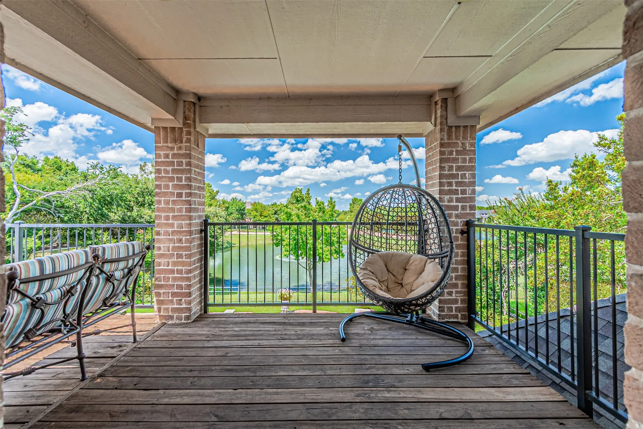 Covered balcony featuring wood-finish flooring, brick columns, and black metal railings overlooking a body of water with surrounding greenery