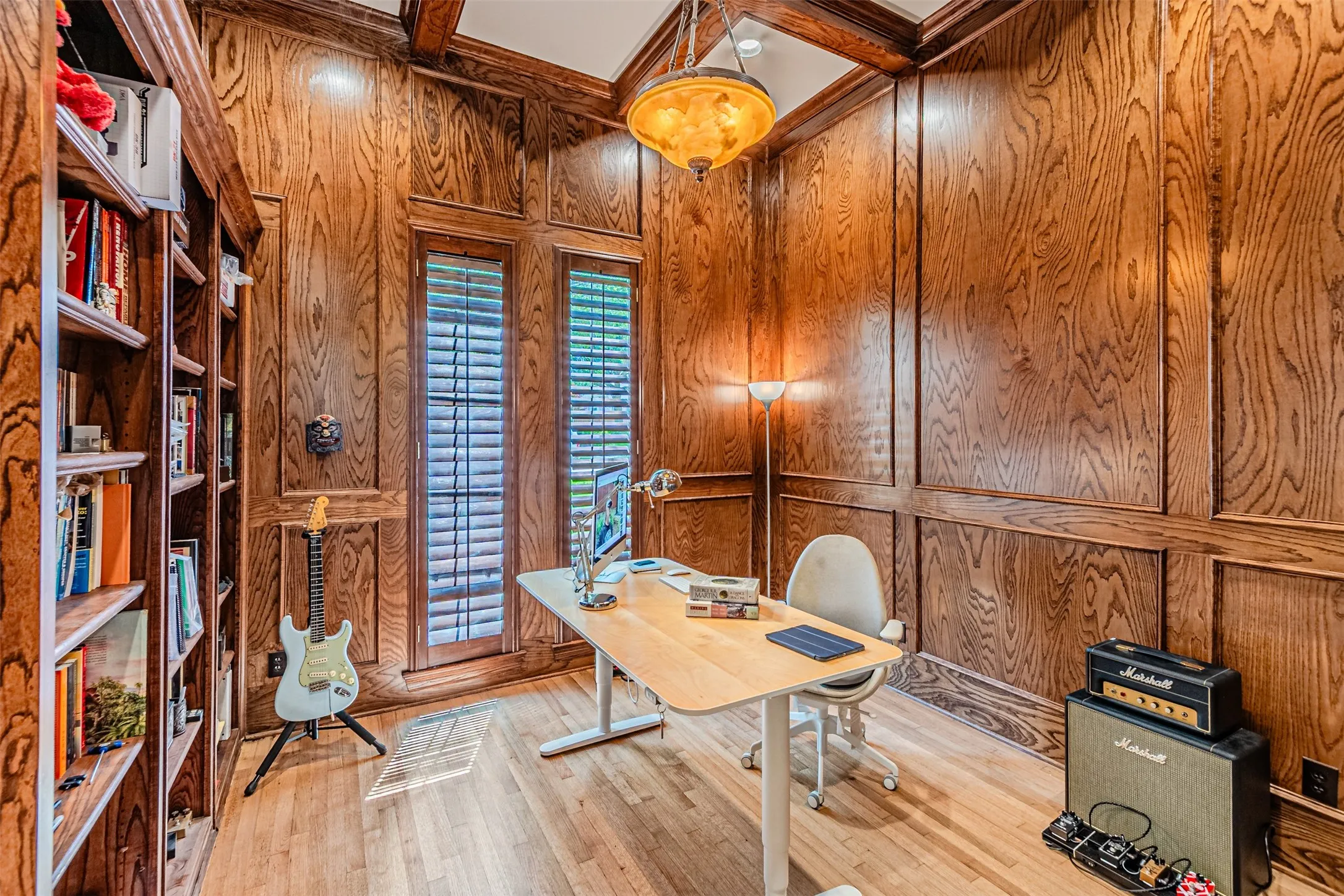 Room featuring extensive wood paneling, wood-finish flooring, a built-in bookshelf, wood-framed windows with plantation shutters, and a coffered ceiling with a central chandelier