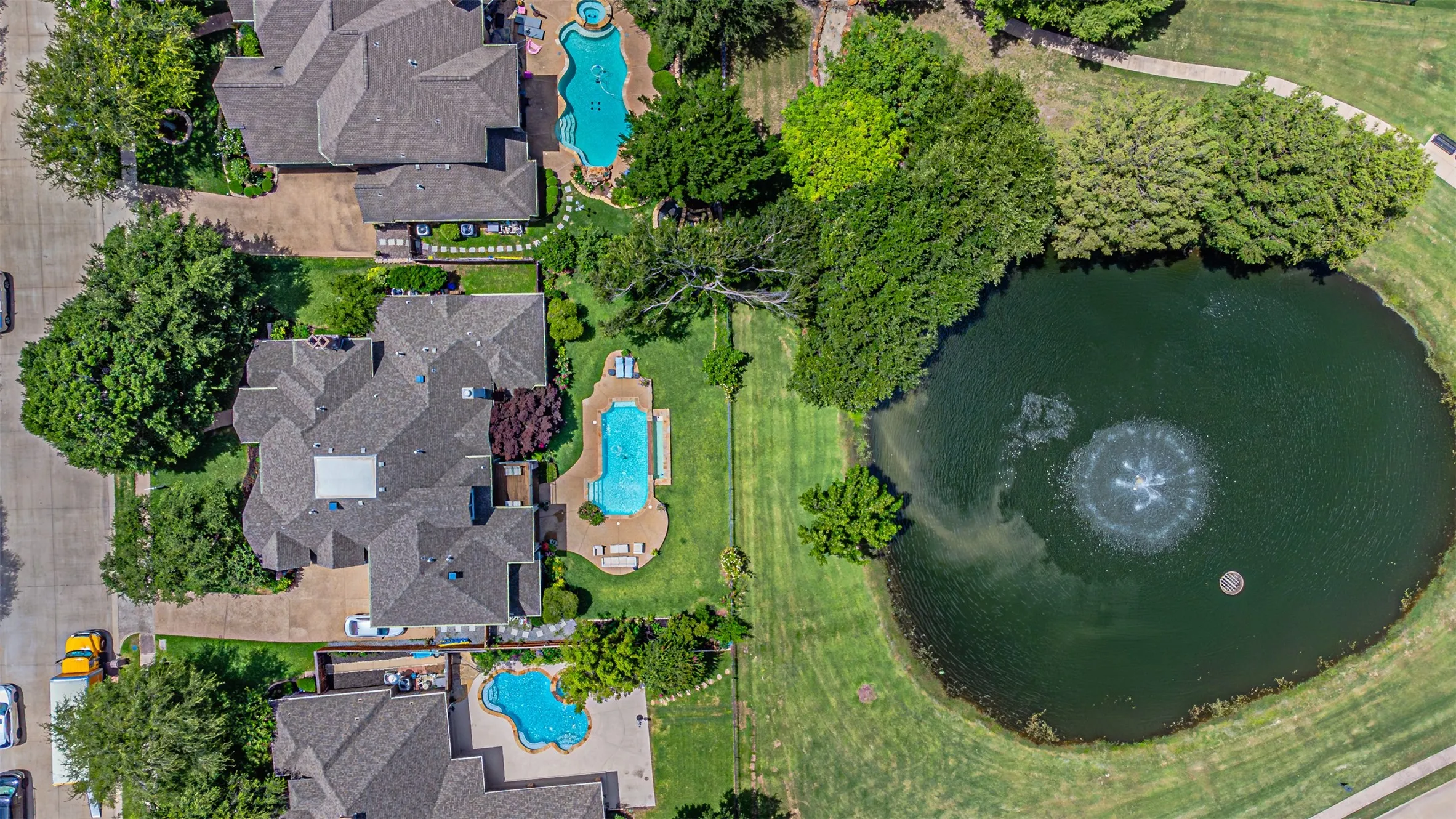 Manicured backyard featuring a freeform swimming pool with integrated spa, extensive hardscaping, and a large grassy area bordering a pond with a fountain