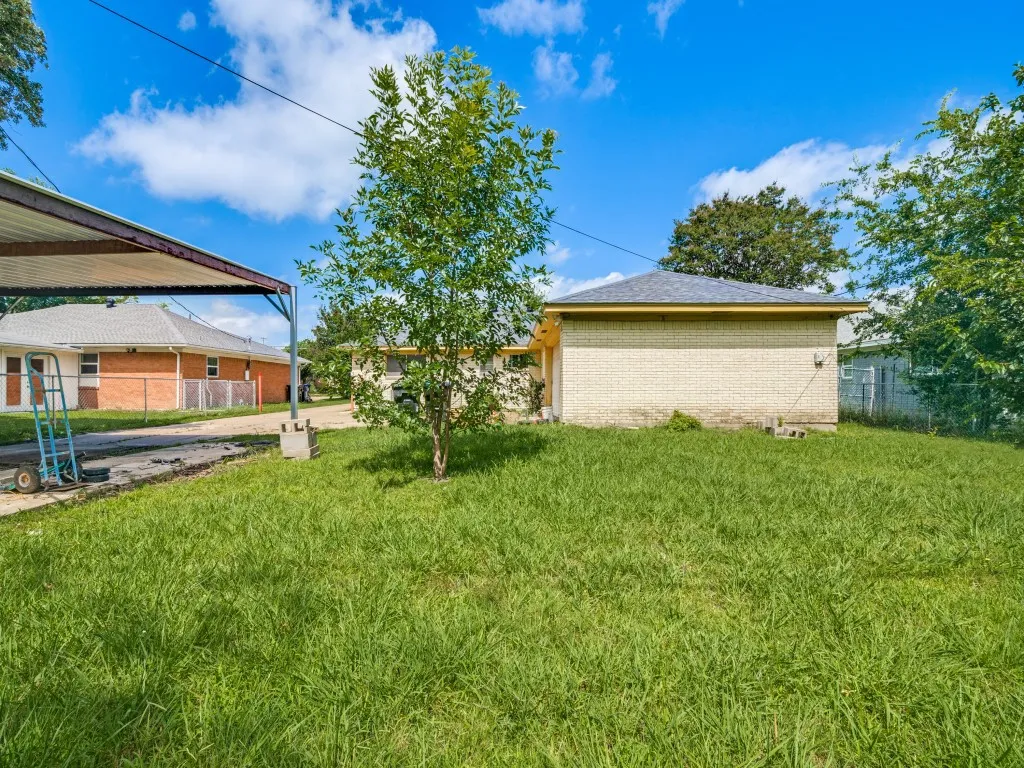 Expansive grassy backyard featuring a partial brick exterior, a covered carport, and mature trees
