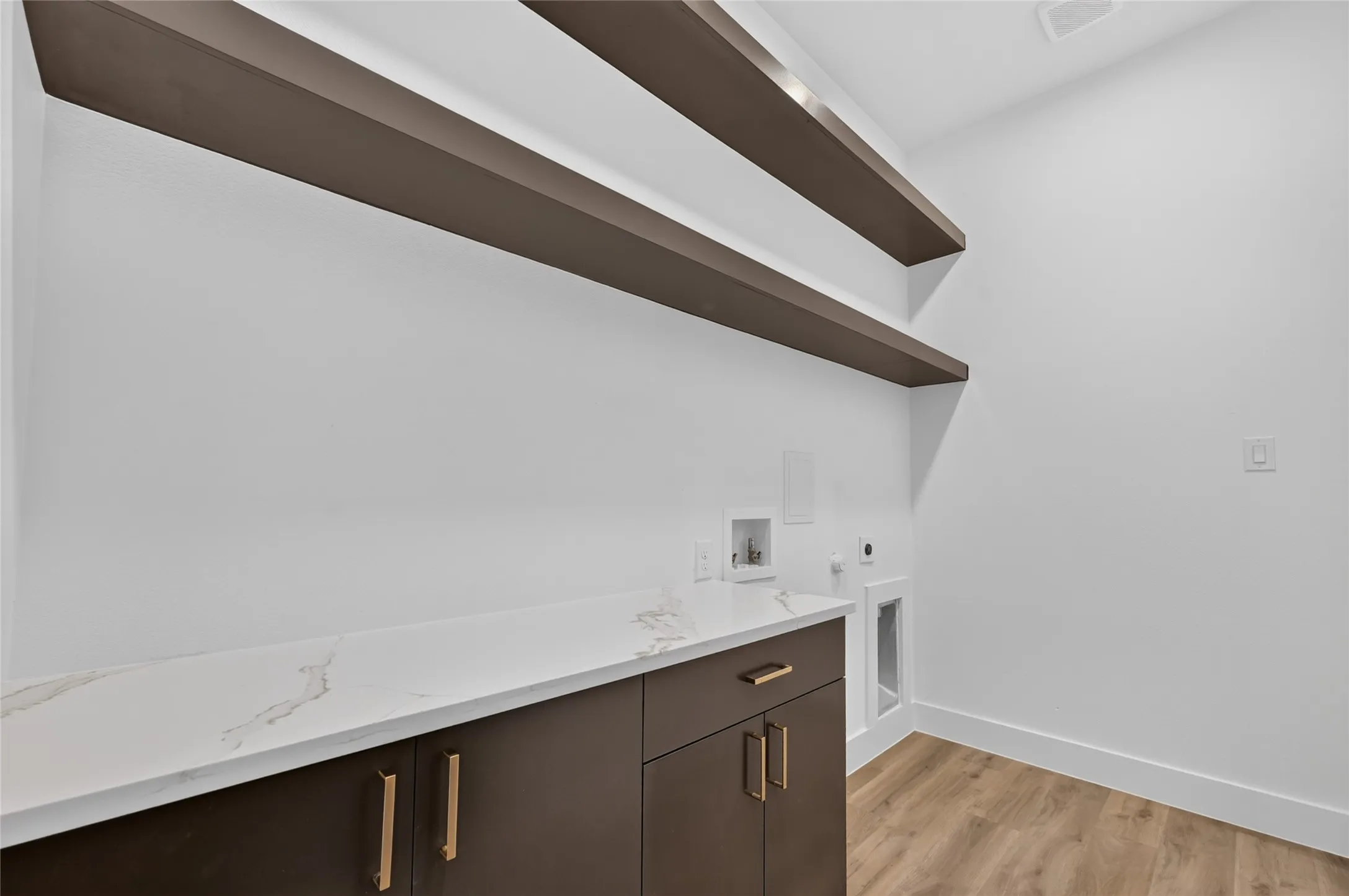 Laundry room featuring a white countertop with subtle veining, dark brown cabinetry with gold hardware, and two long floating shelves