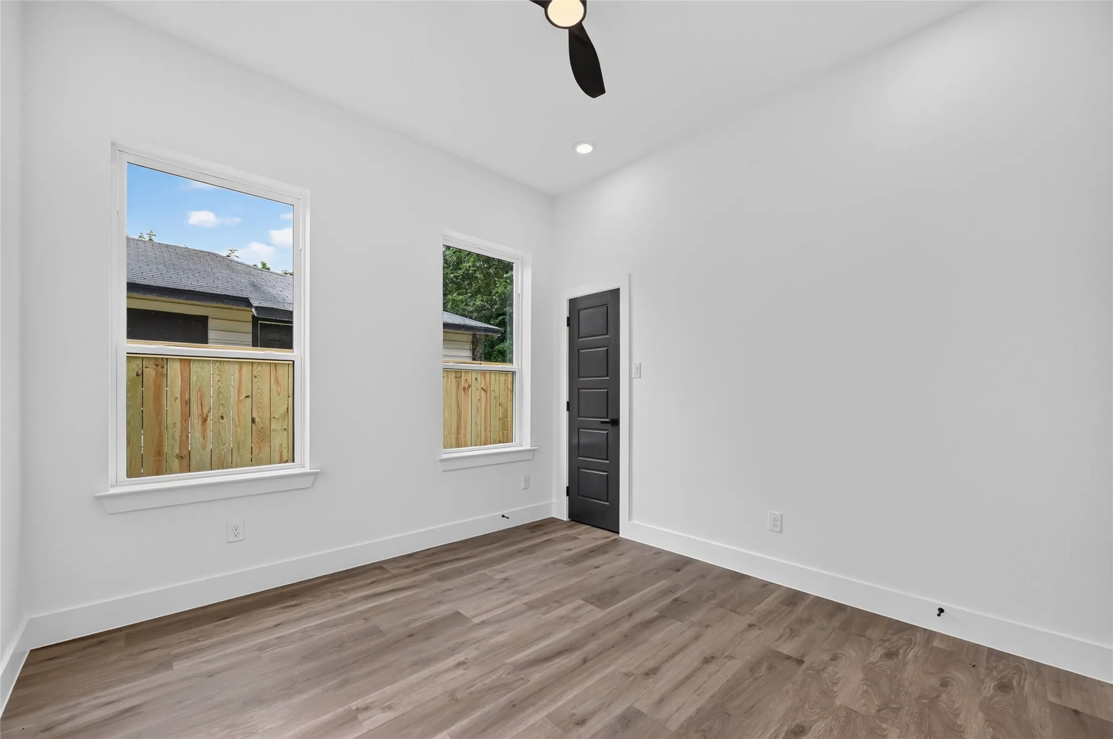Spacious room featuring wood-finish flooring, two large windows, a ceiling fan, recessed lighting, and a modern dark panel door